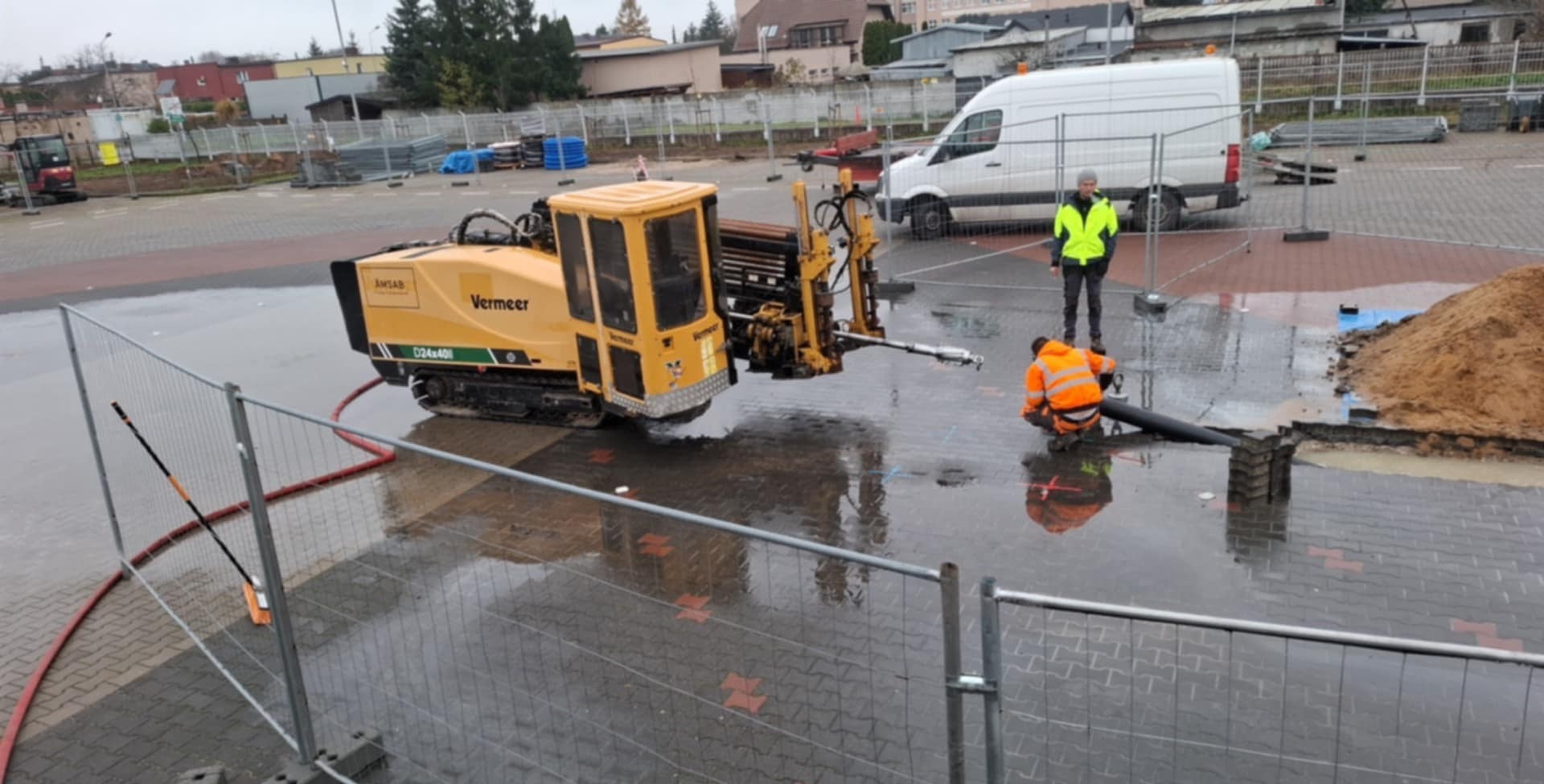 Żółta maszyna Vermeer D24x40II do wiercenia horyzontalnego na mokrym placu budowy, pracownik w pomarańczowej kurtce łączy rury, drugi nadzoruje.