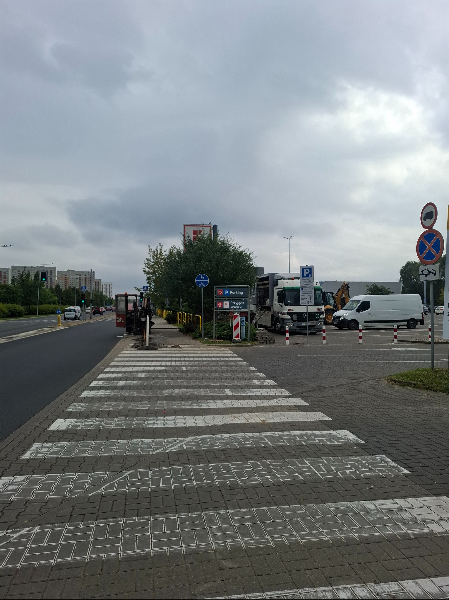 Maszyna do przewiertów przy przejściu dla pieszych, obok zaparkowane busy i koparka. Widok na budynki mieszkalne w tle.