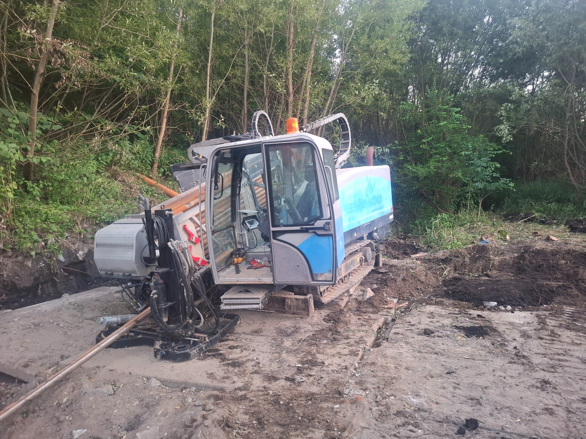 Maszyna do robót ziemnych z otwartą kabiną na placu budowy, w tle zieleń. Widoczne ślady pracy w ziemi.