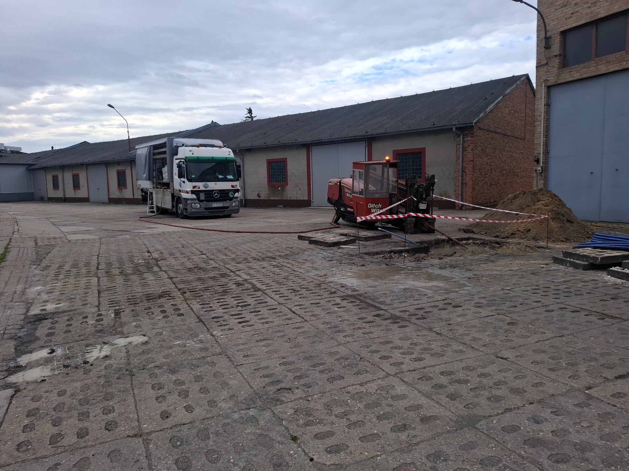 Prace ziemne: wiertnica Ditch Witch przy wykopie zabezpieczonym taśmą, obok ciężarówka z osprzętem na betonowym placu, budynki w tle.