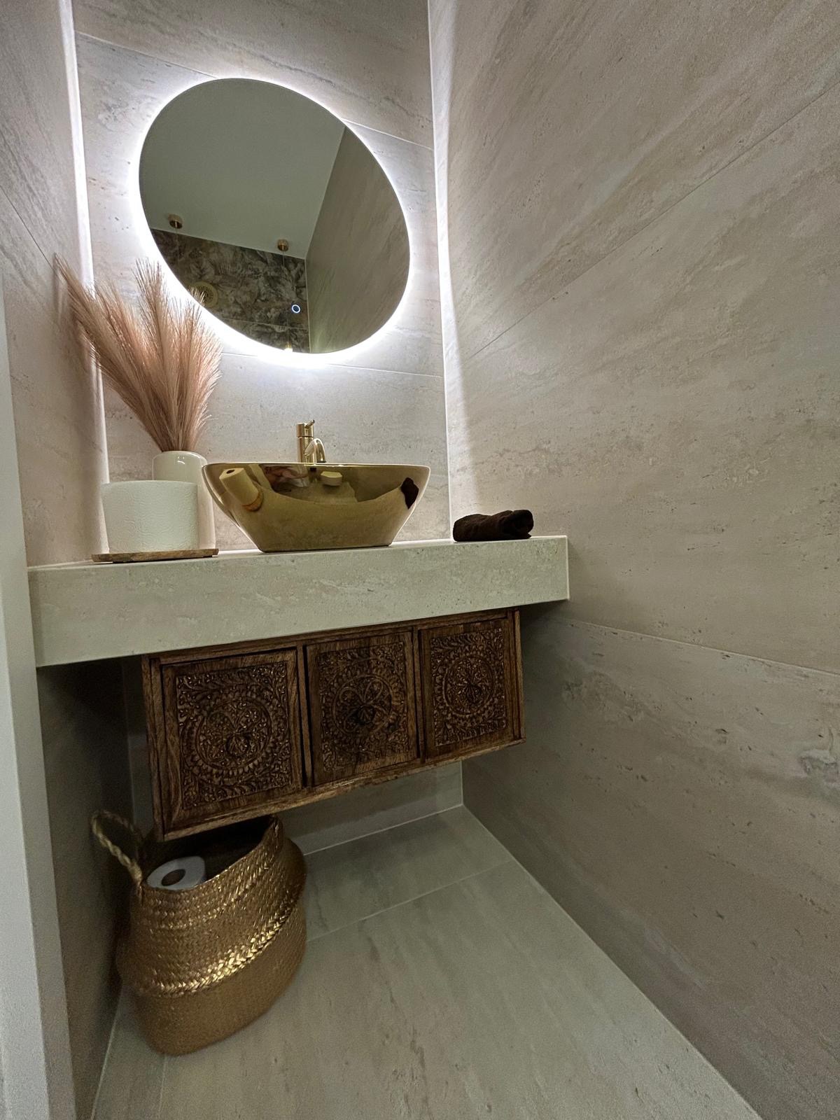 Wykończona łazienka z beżowymi płytkami, złotą umywalką i szafką z rzeźbionymi frontami. Okrągłe lustro z podświetleniem LED. Dekoracyjny kosz i trawa pampasowa.