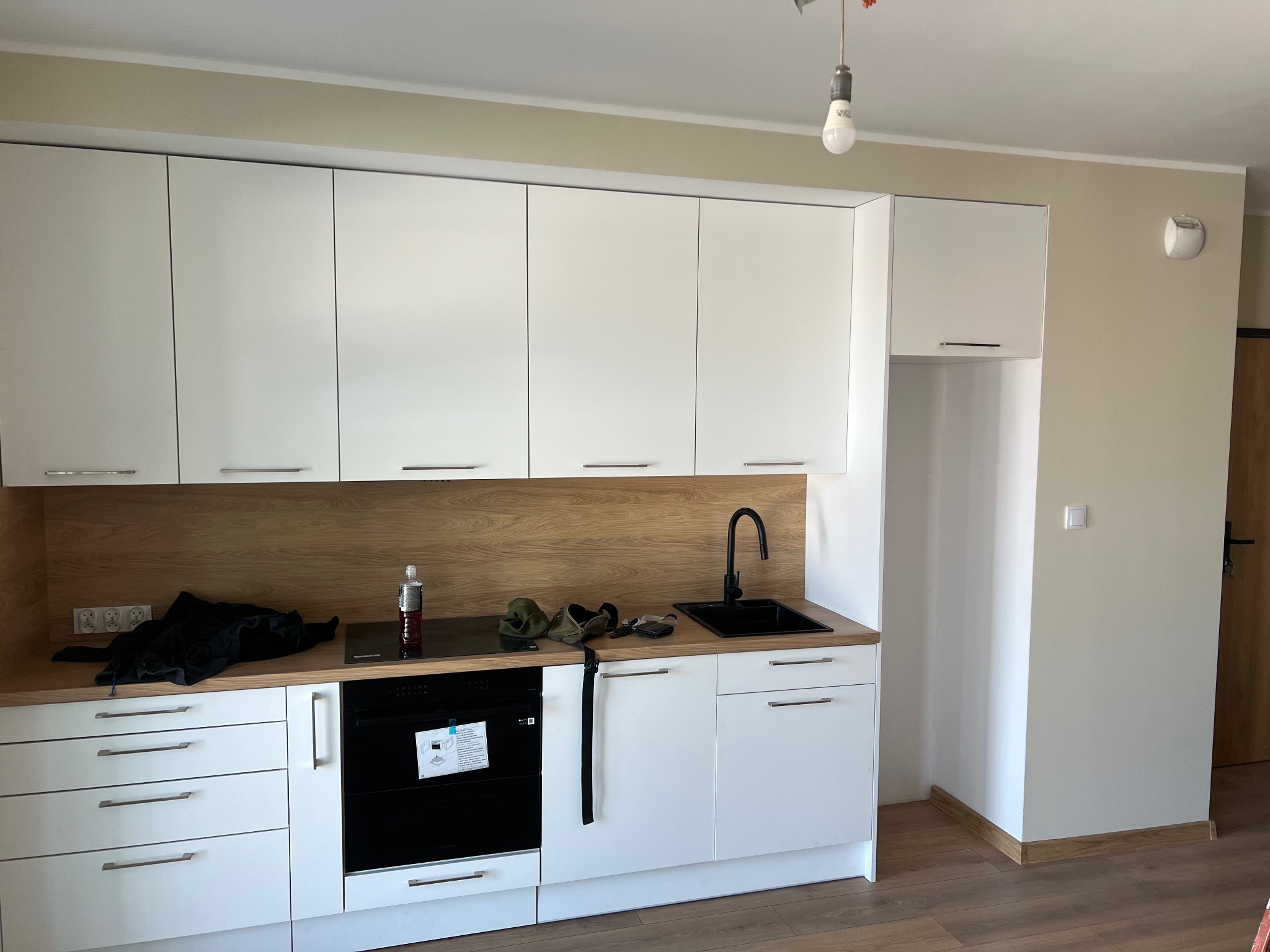 Nowoczesna, jasna kuchnia z białymi szafkami i drewnianym blatem. Czarny zlew i płyta indukcyjna. Widoczne gniazdka elektryczne i akcesoria kuchenne na blacie.