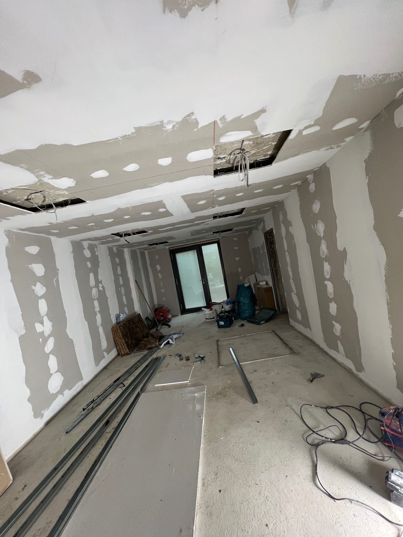 Wnętrze w trakcie prac z płytami karton-gips na ścianach i suficie, widoczne profile, szpachlowanie, kable wystające z otworów w suficie. Prace wykończeniowe w toku.