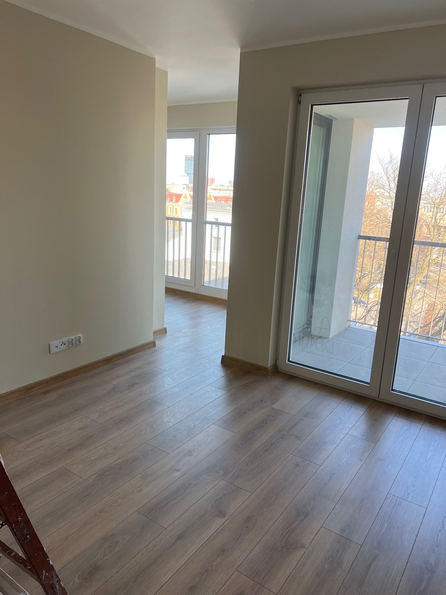 Jasne panele podłogowe w nowym mieszkaniu z balkonem i widokiem na miasto. Świeżo pomalowane ściany i gniazdka elektryczne. Fragment drabiny w lewym dolnym rogu.
