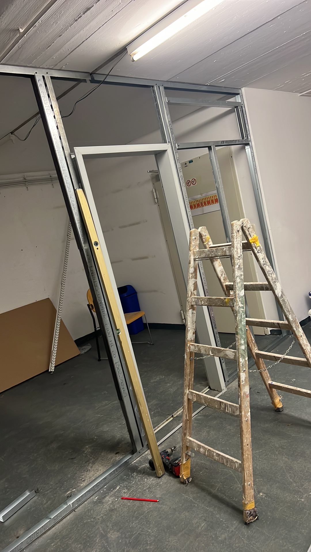 Montaż ścianki działowej z profili metalowych, widoczna poziomica, wiertarka i drabina malarska. Prace wykończeniowe w pomieszczeniu z surowym oświetleniem.