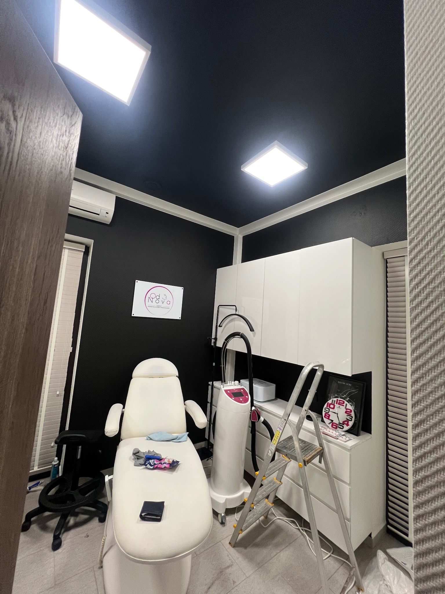 Nowoczesne wnętrze gabinetu kosmetycznego z czarnymi ścianami i sufitem, białymi meblami, fotelem zabiegowym i lampami sufitowymi. Widoczna drabina i sprzęt kosmetyczny.