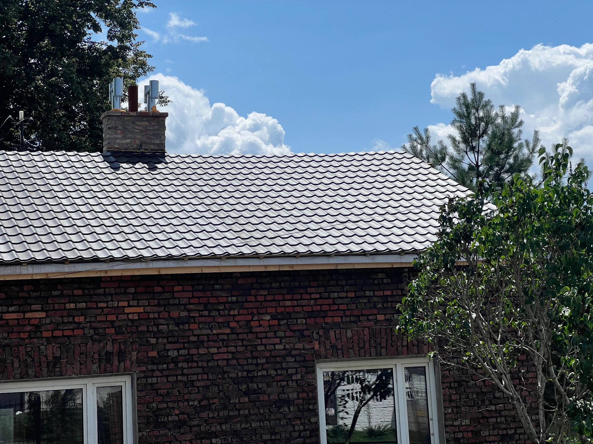 Ceglany dom z nowym, błyszczącym dachem w odcieniach szarości, komin z metalowymi wkładami, fragment okna i zieleni na tle błękitnego nieba z chmurami.