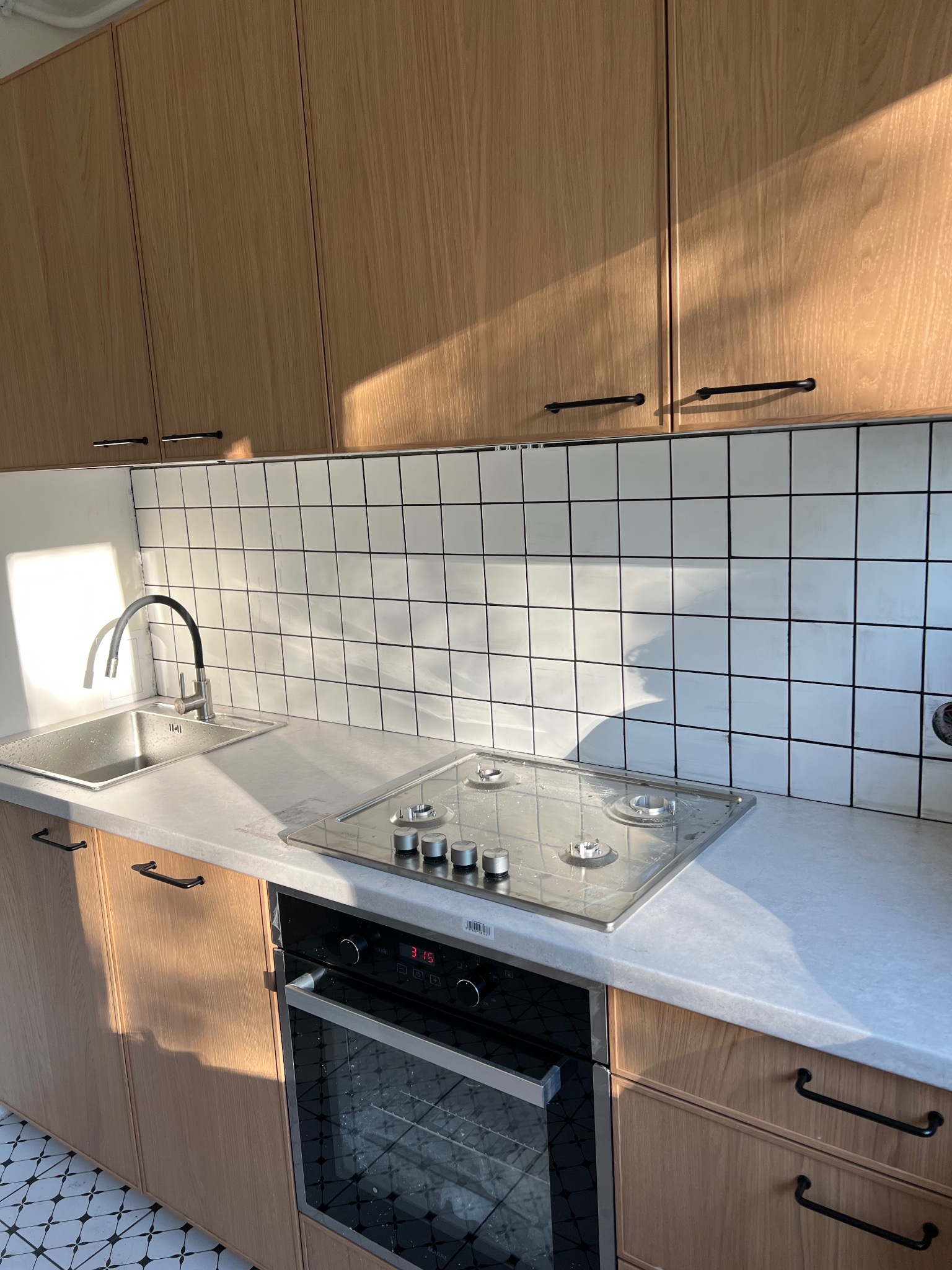 Jasna kuchnia z drewnianymi szafkami, białymi płytkami i czarnymi uchwytami. Ujęcie na zlew, płytę gazową i piekarnik. Na podłodze wzorzyste płytki.