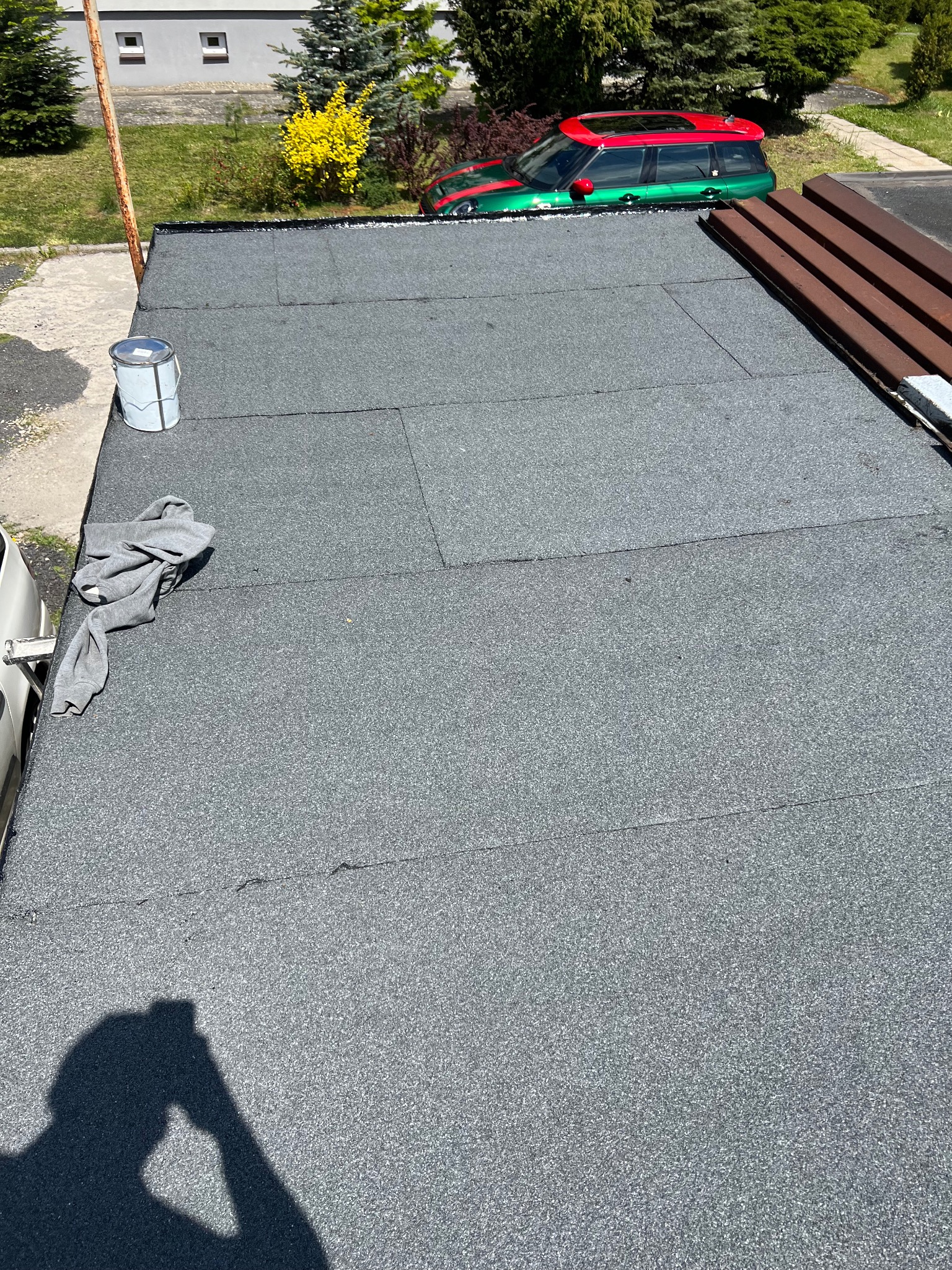 Szary dach pokryty papą termozgrzewalną, widoczny z góry, z elementami wykończeniowymi i cieniem osoby robiącej zdjęcie. W tle zieleń i budynek.