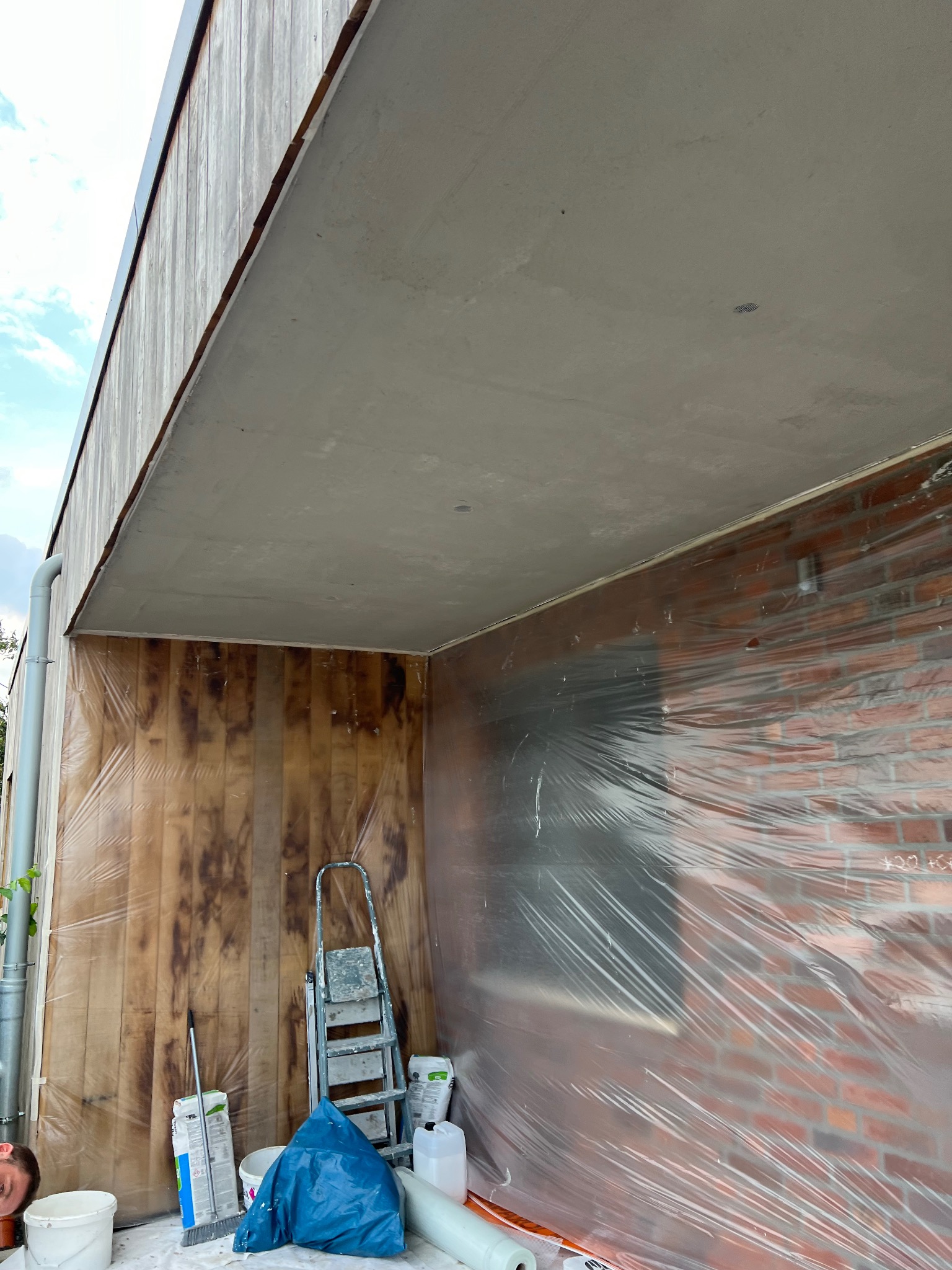 Przygotowania do prac wykończeniowych: zabezpieczona folią ściana z cegły, drewniana boazeria, drabina i materiały budowlane w pomieszczeniu przed malowaniem sufitu.