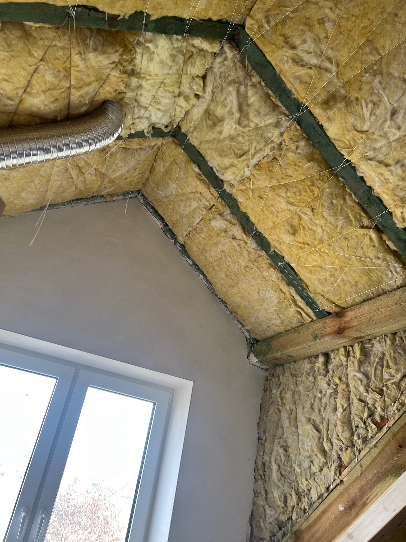 Ocieplenie poddasza wełną mineralną, widoczne drewniane elementy konstrukcyjne, fragment okna i rura wentylacyjna. Prace wykończeniowe w trakcie realizacji.