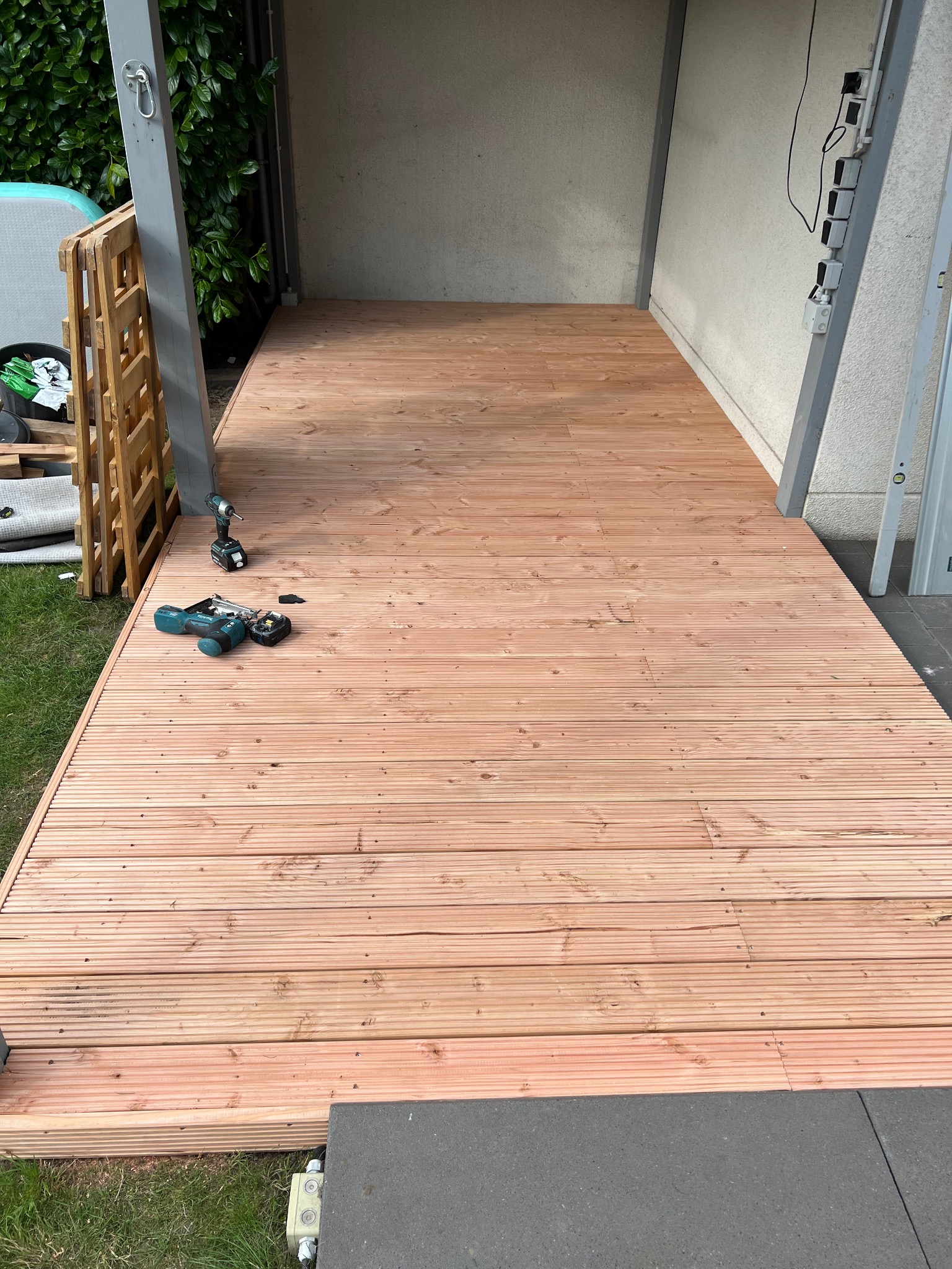 Nowa, jasna drewniana podłoga tarasu z widocznymi narzędziami pracy, częściowo na zewnątrz budynku. Widoczne deski z wyraźnym usłojeniem i wkręty mocujące.
