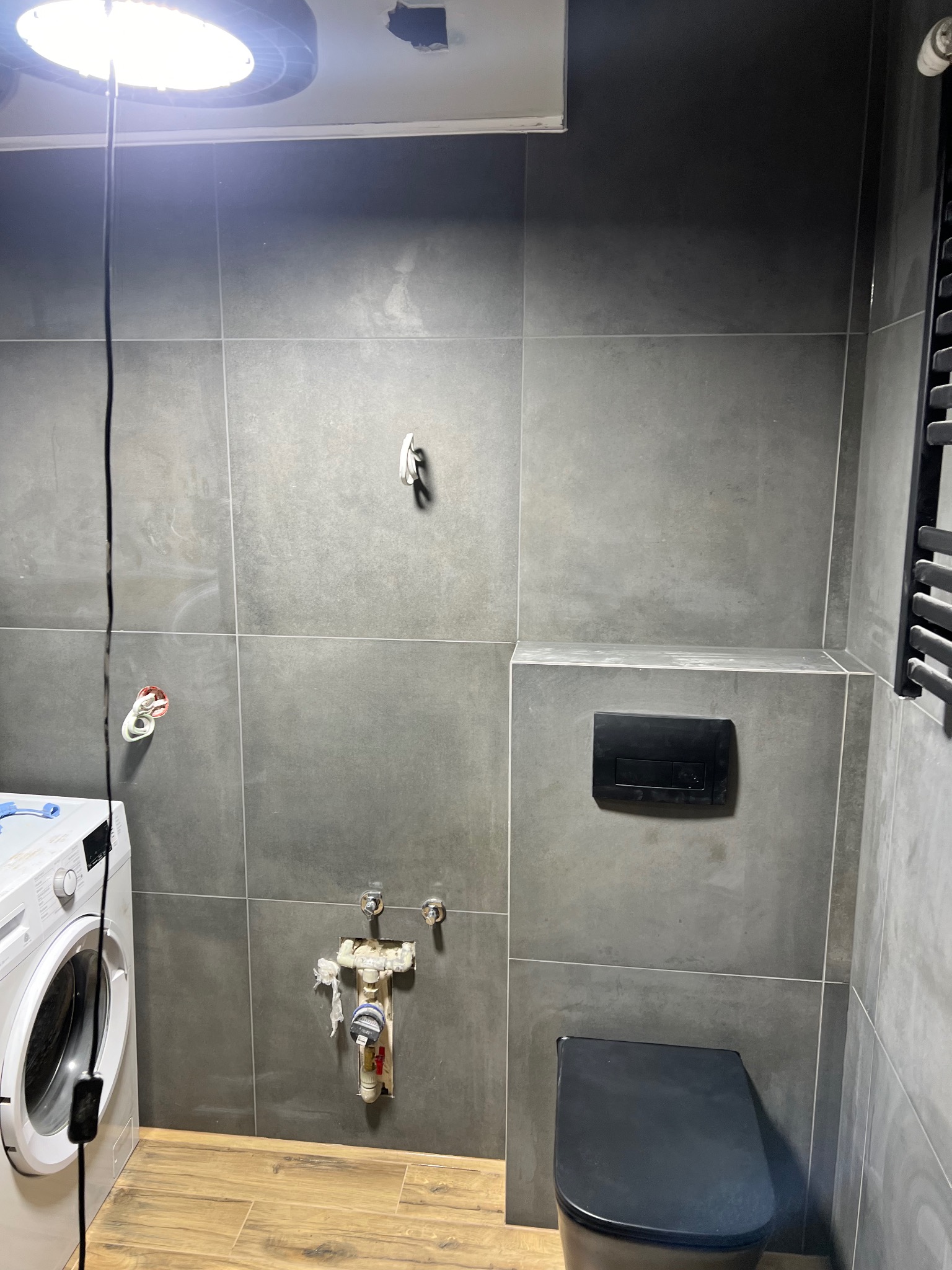 Szara łazienka w trakcie remontu: widoczna pralka, toaleta, grzejnik drabinkowy i instalacja wodna z zabezpieczeniami. Podłoga wyłożona panelami imitującymi drewno.