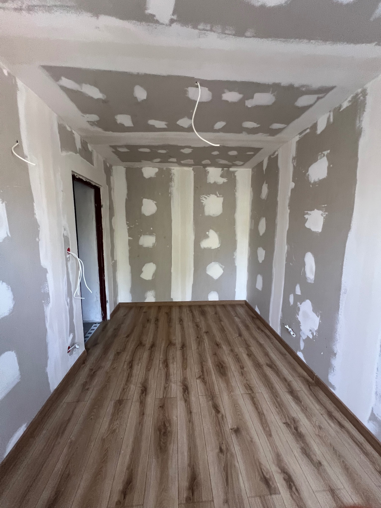 Pokój w trakcie wykończenia: ułożona podłoga z paneli, ściany z płyt gipsowo-kartonowych z widocznymi łączeniami i przewodami elektrycznymi. Widok na surowe wnętrze.