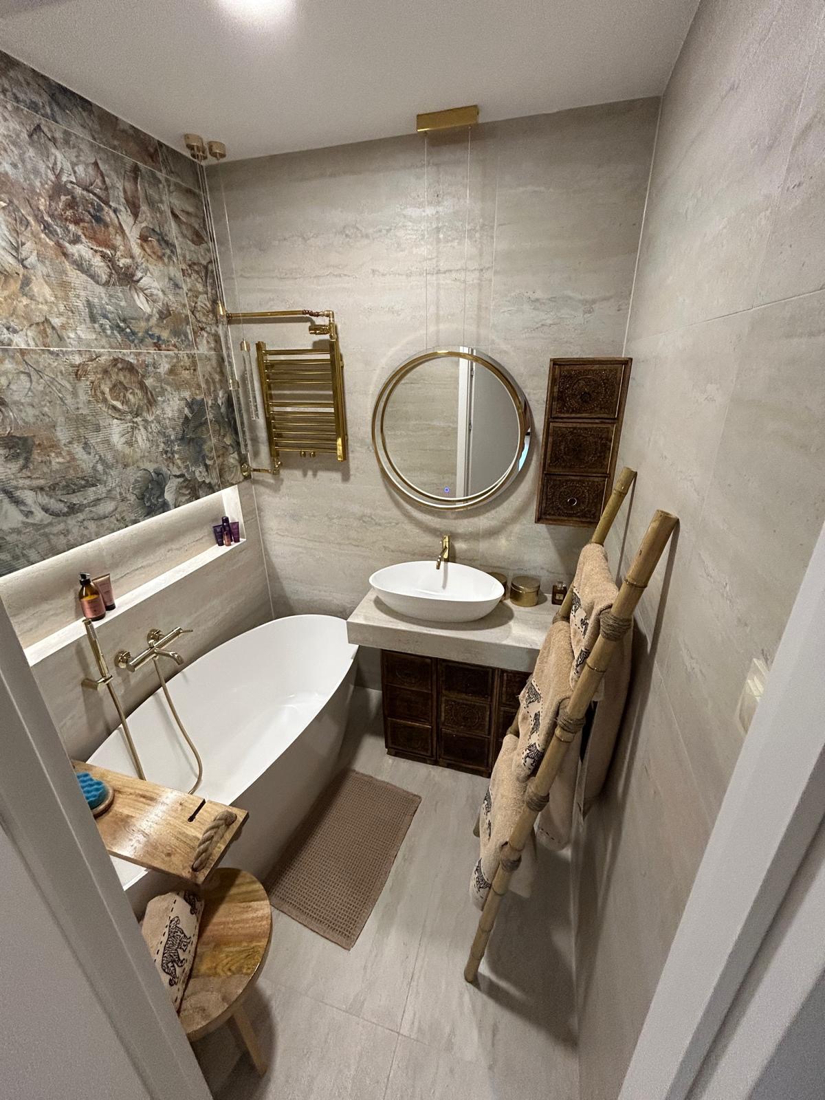 Stylowa łazienka z wanną, umywalką na szafce z rzeźbionymi drzwiczkami i okrągłym lustrem w złotej ramie. Na ścianie dekoracyjna płytka i grzejnik drabinkowy.