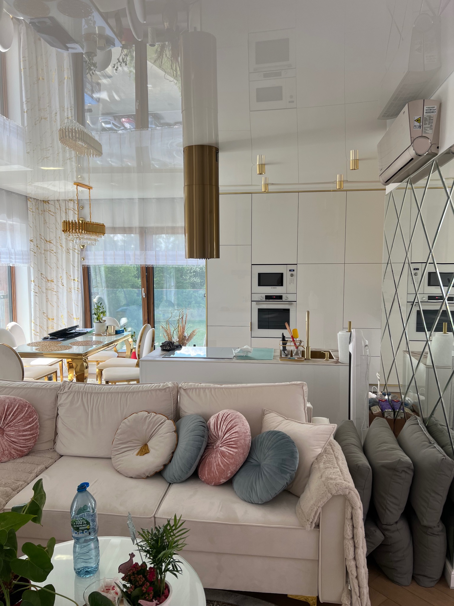 Eleganckie wykończenie wnętrza w Katowicach: jasna kuchnia z wyspą, lustrzana ściana i wygodna sofa z dekoracyjnymi poduszkami. Nowoczesny design i dbałość o detale.