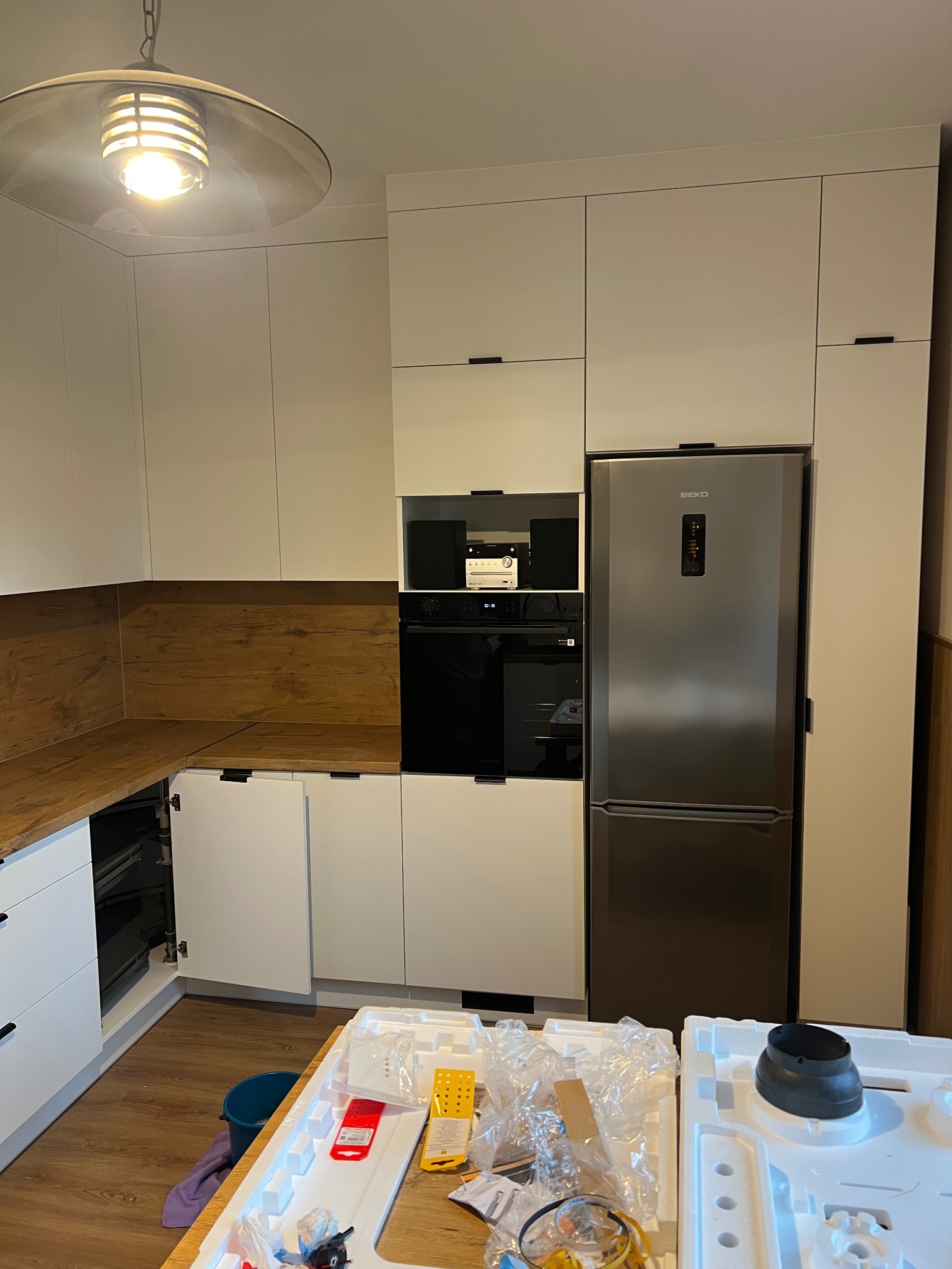 Nowoczesna kuchnia z białymi szafkami, drewnianym blatem i srebrną lodówką Beko. Na pierwszym planie elementy montażowe, sugerujące etap wykończenia.