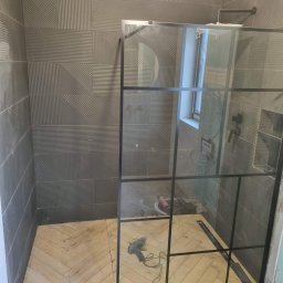 Firma Remontowo Budowlana - Nowoczesna łazienka w trakcie wykańczania: szare płytki z wzorem, drewnopodobna podłoga w jodełkę, czarna kabina prysznicowa i narzędzia glazurnicze.