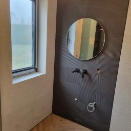 Firma Remontowo Budowlana - Nowoczesna łazienka w trakcie wykańczania: ciemne, wzorzyste płytki, okrągłe lustro, drewniana podłoga w jodełkę, widoczne elementy instalacji. Minimalistyczny design.