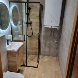 Firma Remontowo Budowlana - Nowoczesna łazienka po remoncie: kabina prysznicowa typu walk-in z czarnymi ramami, umywalka z szafką w odcieniach drewna, okrągłe lustro z podświetleniem LED i bojler.