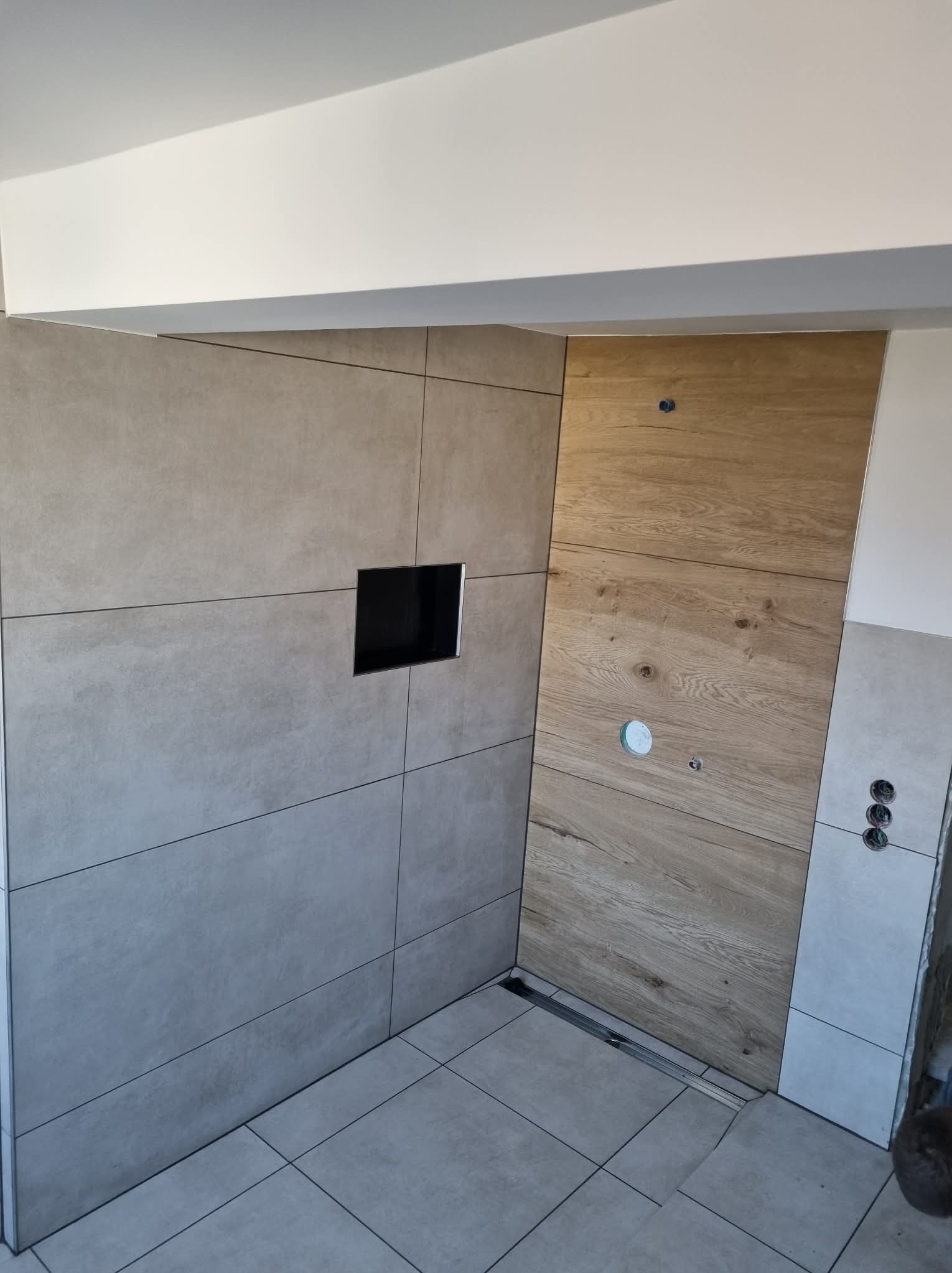 Nowoczesna łazienka w trakcie wykańczania: szare płytki wielkoformatowe, drewnopodobne panele ścienne, odpływ liniowy. Widoczne otwory na armaturę.
