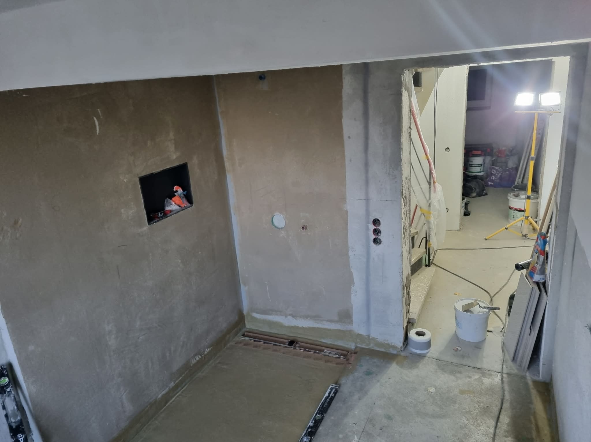 Surowa łazienka w trakcie szpachlowania: ściany pokryte gładzią, widoczny odpływ liniowy w podłodze, narzędzia i materiały budowlane w tle, surowy stan wykończenia.