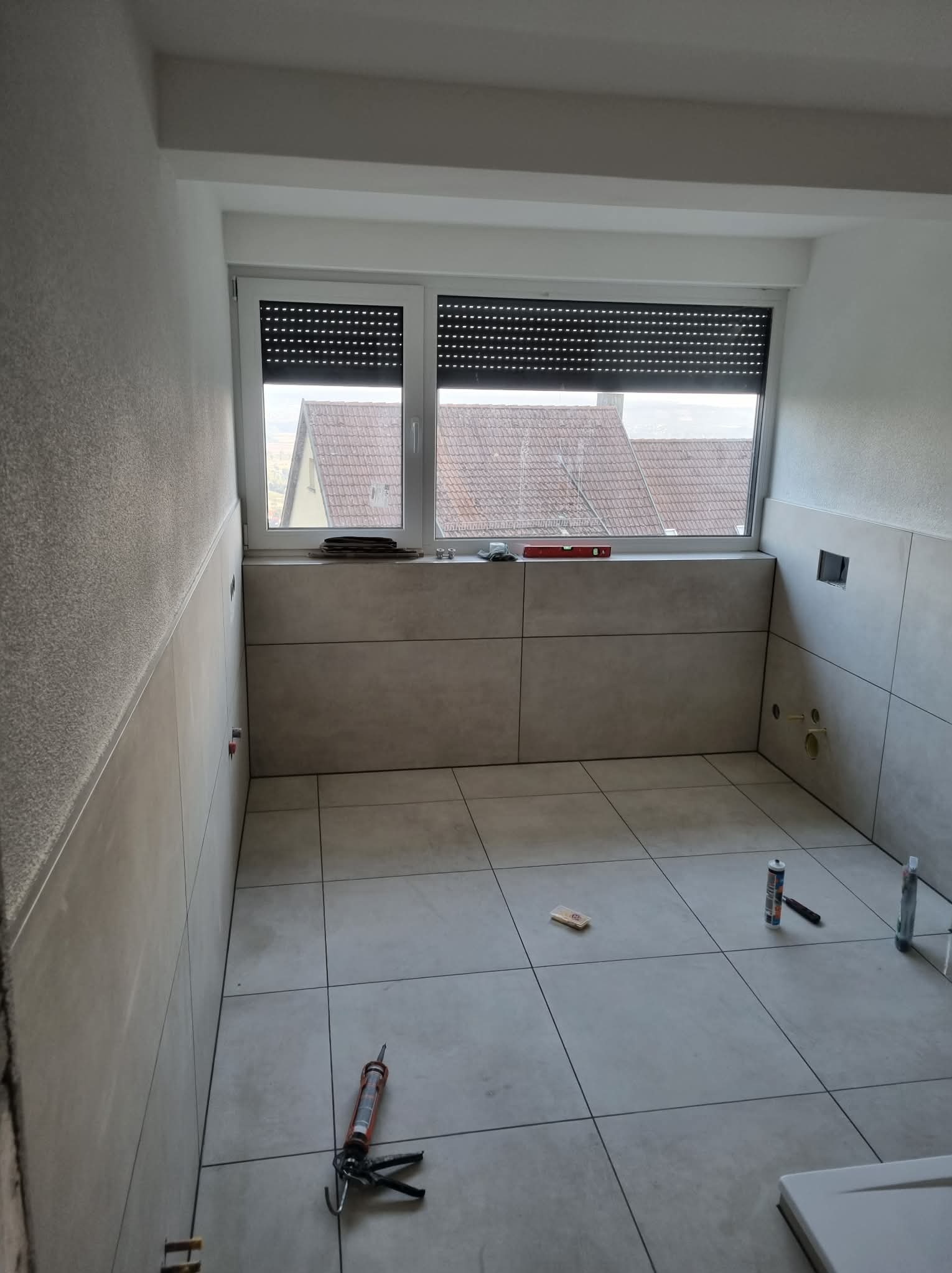 Jasne płytki na ścianach i podłodze w trakcie montażu w pomieszczeniu z oknem. Widoczne narzędzia glazurnicze i poziomica. Ujęcie z góry, neutralne oświetlenie.