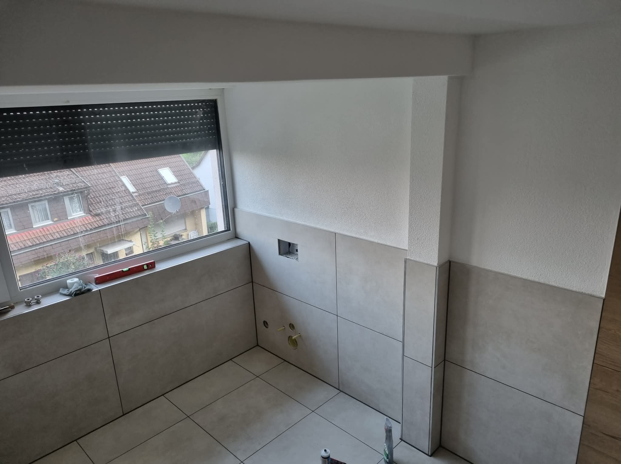 Jasne, kwadratowe płytki częściowo ułożone na ścianach i podłodze w łazience. Widoczne otwory na instalacje i okno z roletą. Poziomica na parapecie.