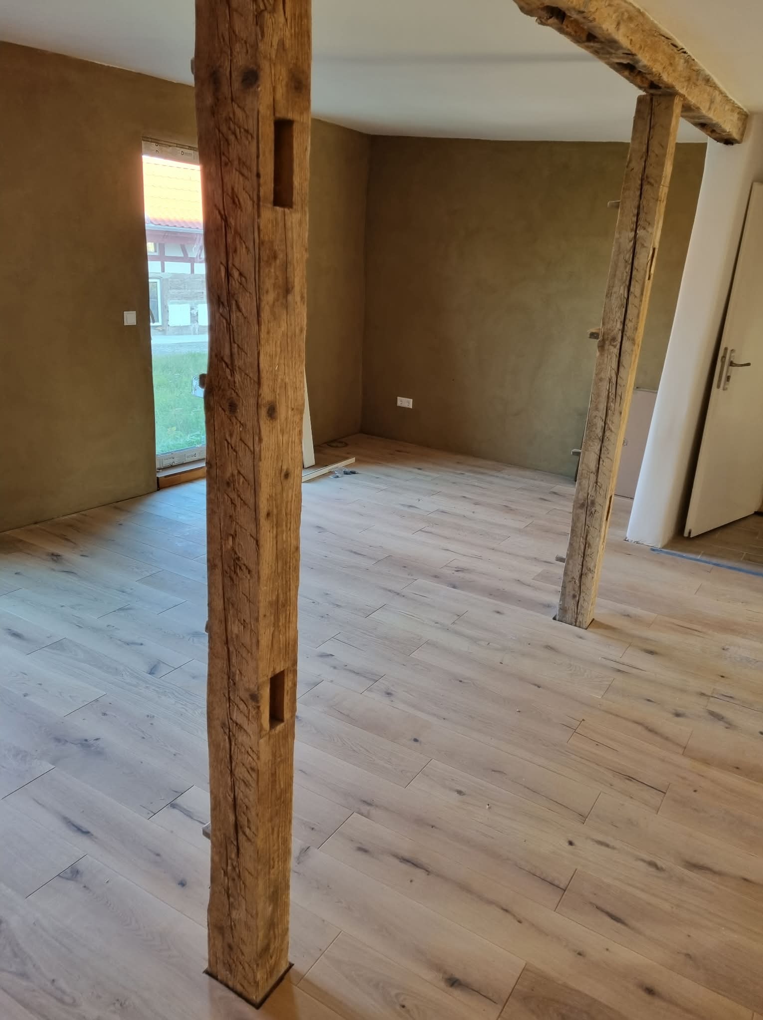 Jasna drewniana podłoga w pokoju z beżowymi ścianami i drewnianymi belkami. Widoczne fragmenty okna i drzwi. Belki z wyraźną strukturą drewna.