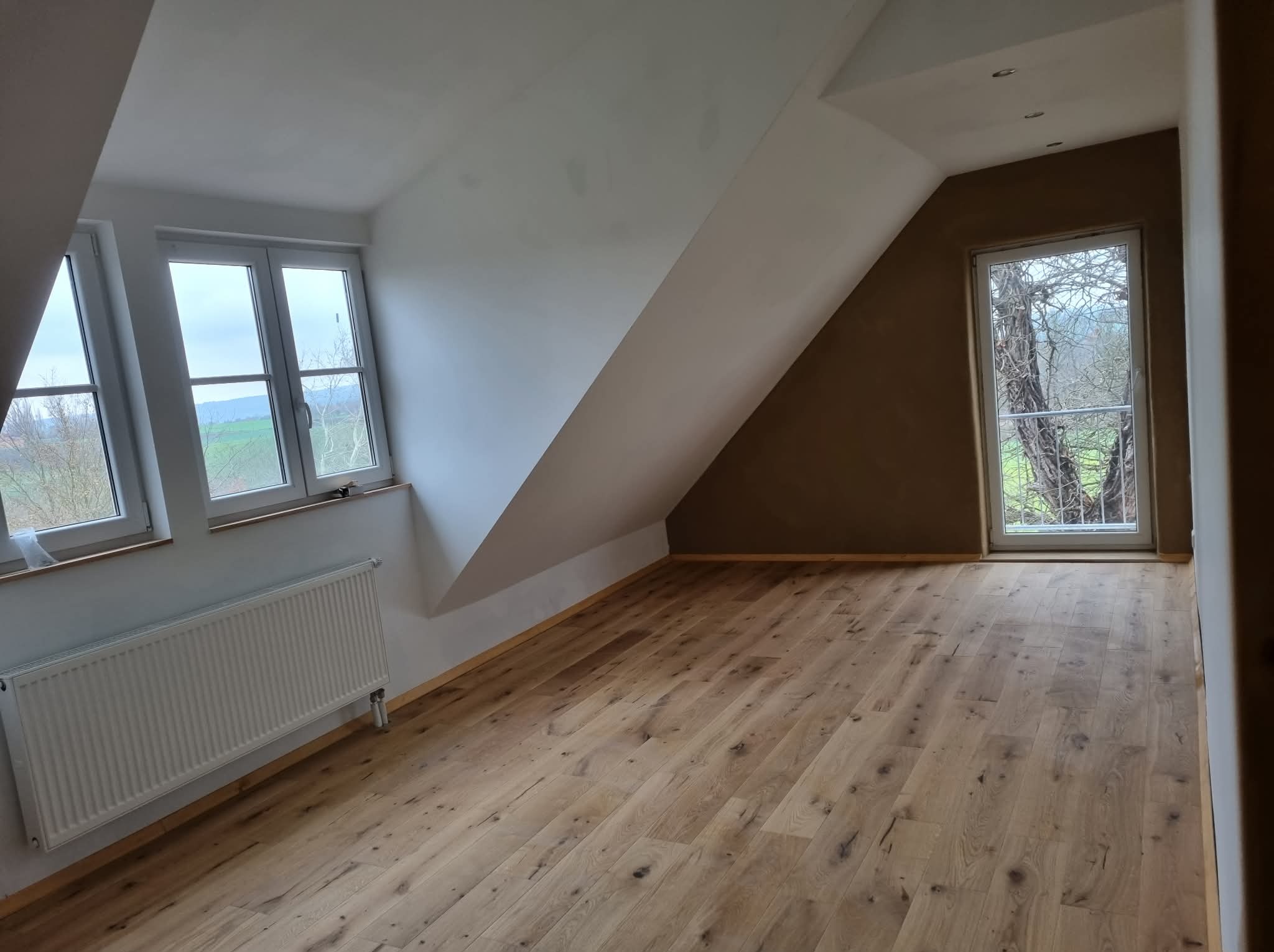 Jasna drewniana podłoga w nowym pokoju na poddaszu z oknami i grzejnikiem. Ściany białe i brązowe. Widok na zewnątrz przez okna.