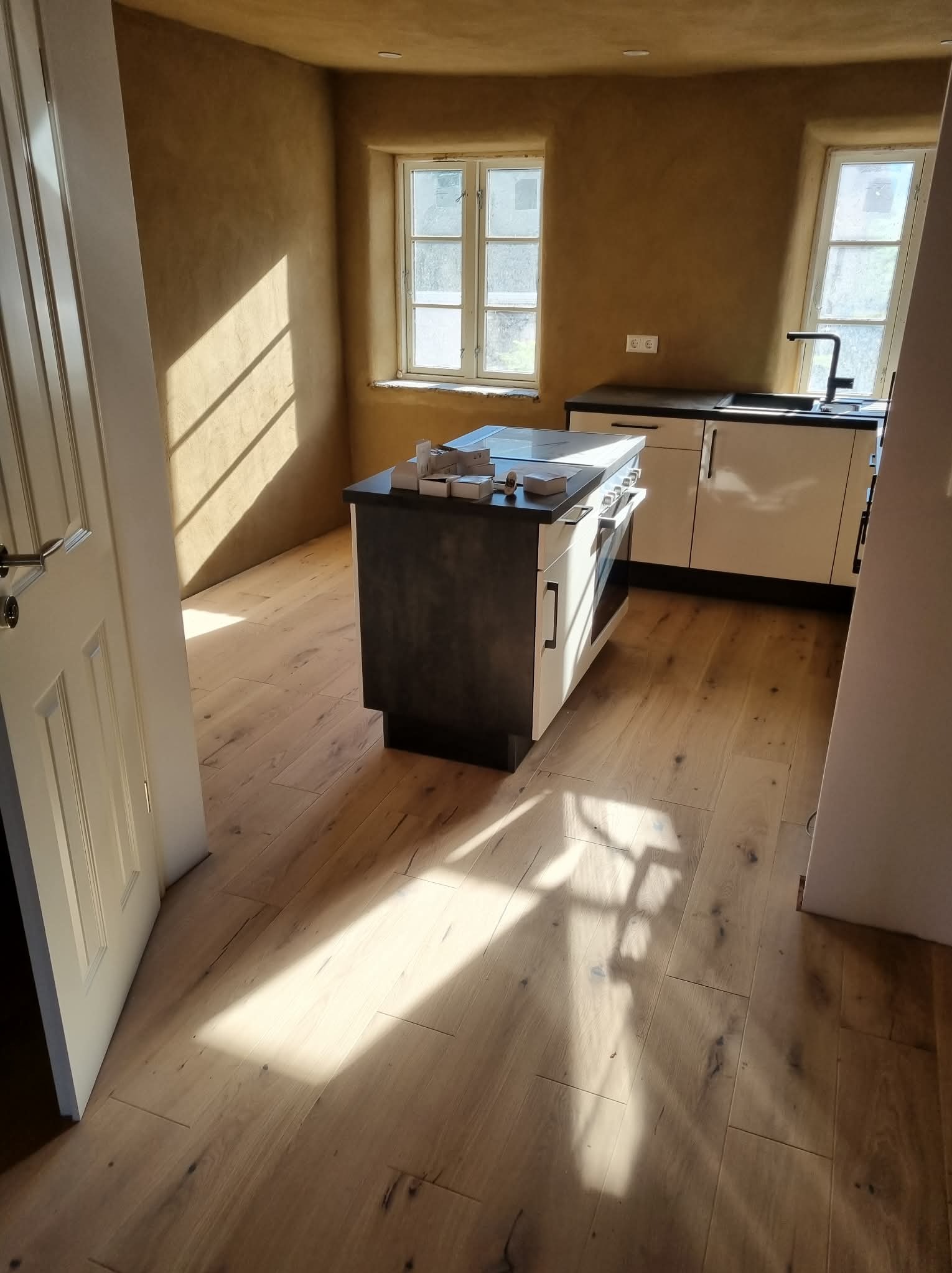 Jasna kuchnia z wyspą i drewnianą podłogą, ściany w odcieniach beżu, wpadające promienie słoneczne przez okna. Widok na wykończone wnętrze.