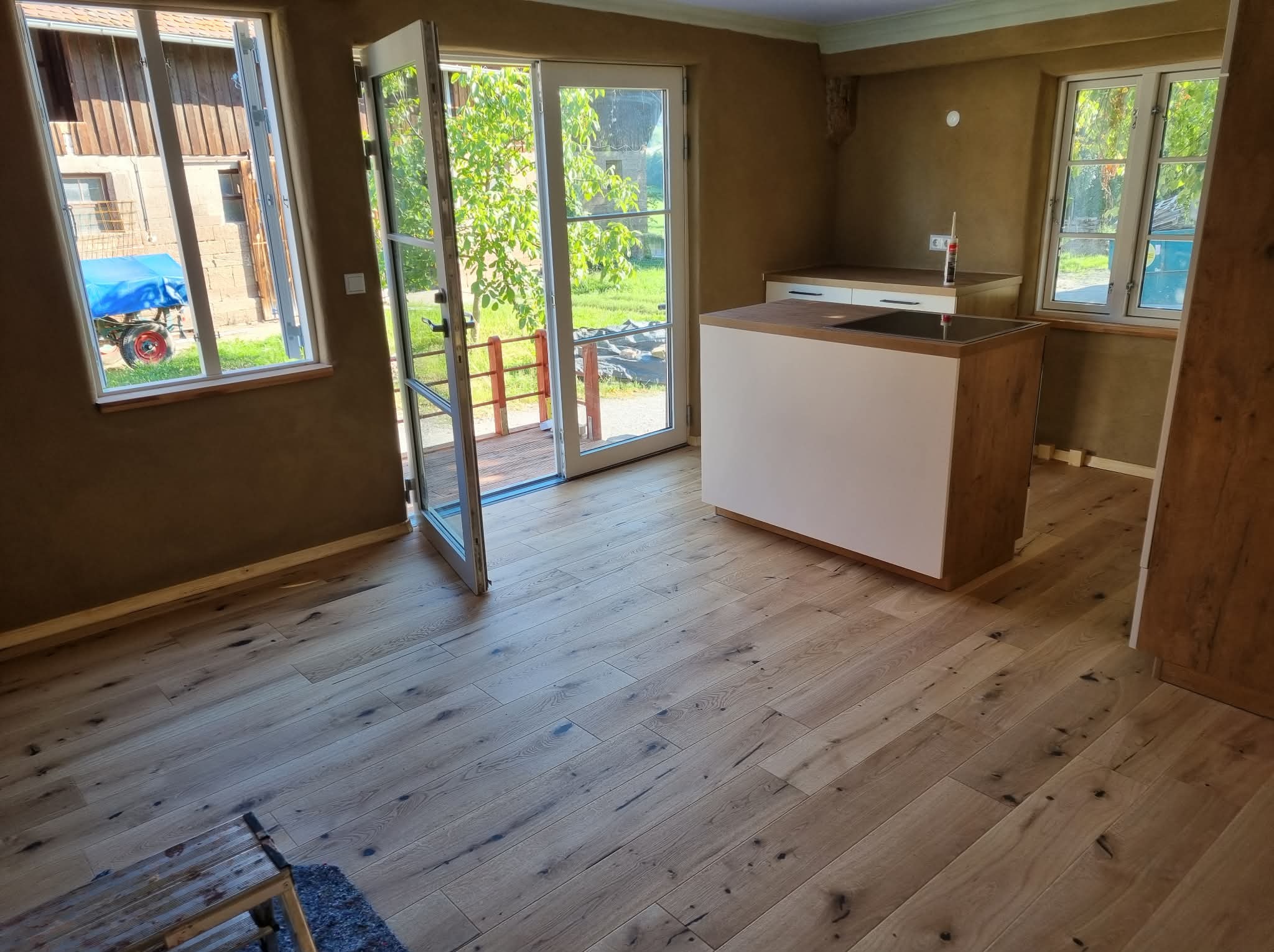 Jasna, drewniana podłoga w nowym wnętrzu z otwartymi drzwiami balkonowymi i oknami. Widoczna wyspa kuchenna i fragment zabudowy. Ściany w odcieniach beżu.