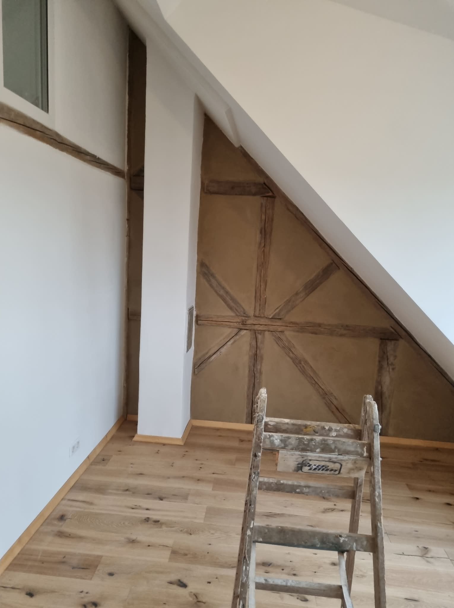 Poddasze po remoncie: białe ściany, drewniana konstrukcja szkieletowa, jasna podłoga i drabina malarska. Widok na wykończone wnętrze z bliska.