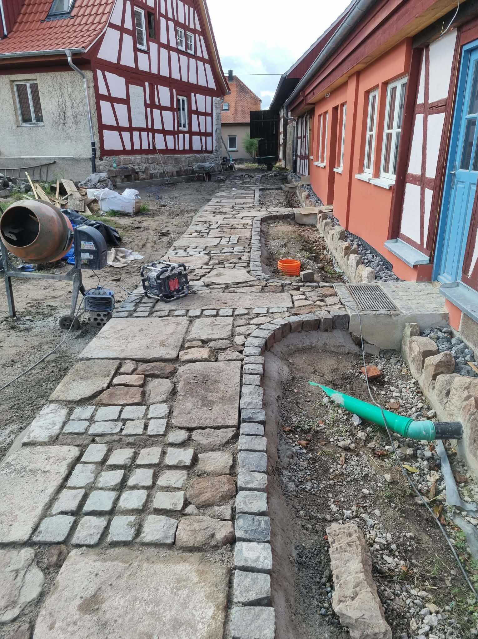 Układanie kamiennej ścieżki z kostki brukowej przy budynku z czerwonymi elementami. Widoczna betoniarka i narzędzia. Etap prac ziemnych i układania obrzeży.