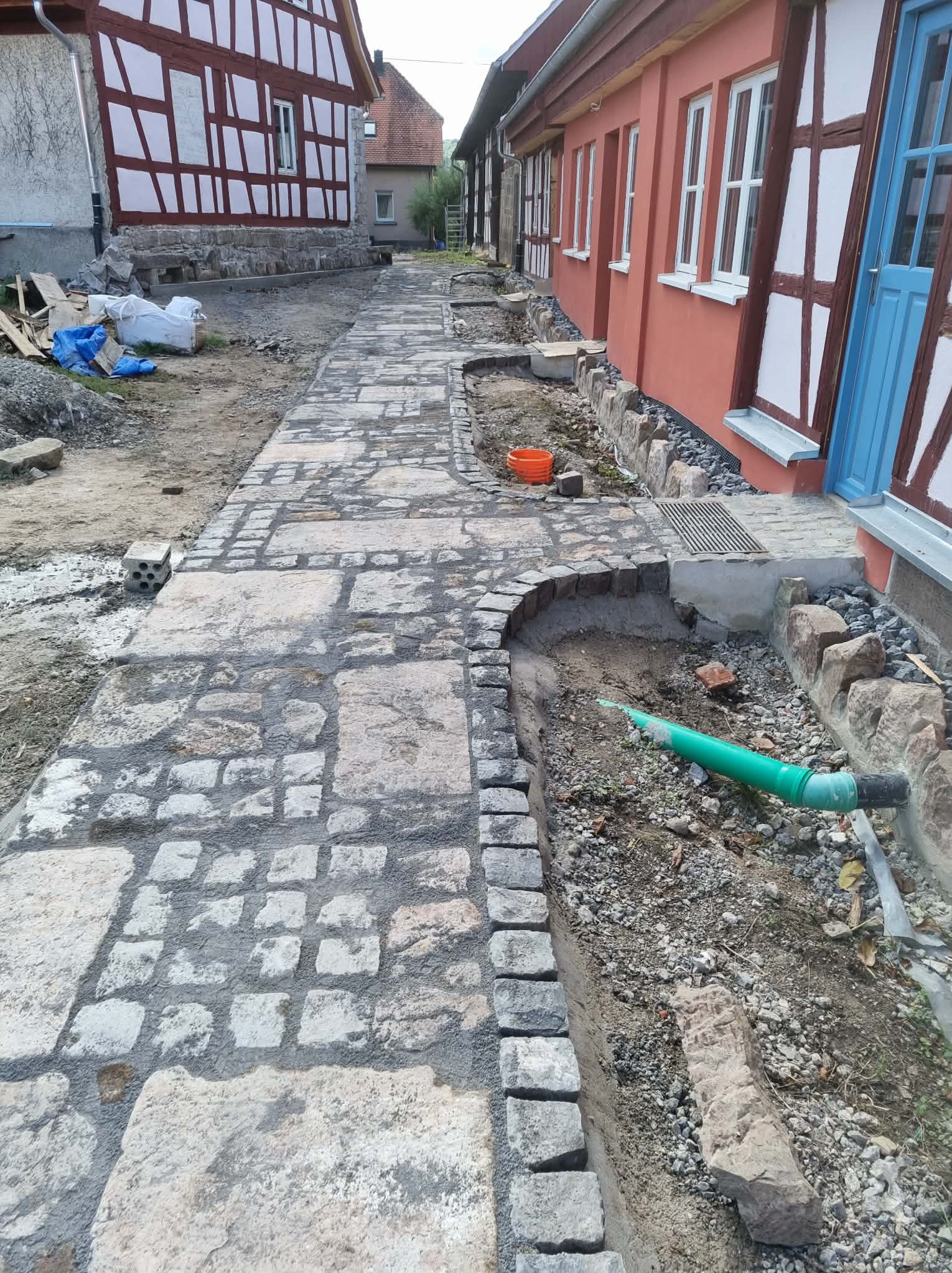 Układanie kamiennej ścieżki wzdłuż budynku z muru pruskiego. Częściowo ułożona kostka, widoczne prace ziemne i instalacja kanalizacyjna. Etap prac wykończeniowych.