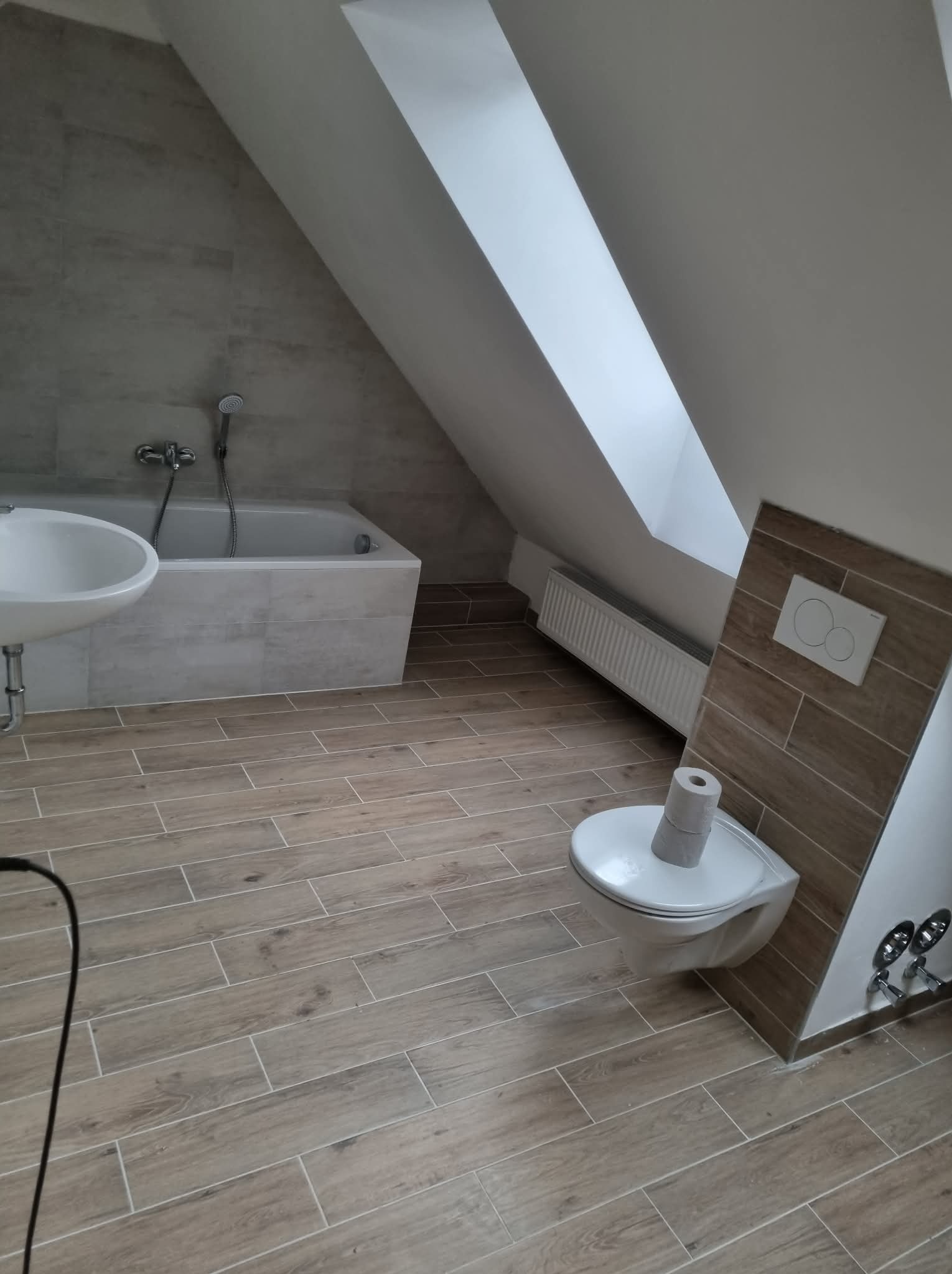 Ukończona łazienka na poddaszu: beżowe płytki imitujące drewno na podłodze i ścianach, biała wanna, umywalka i toaleta pod skosem dachu. Nowoczesne wykończenie.