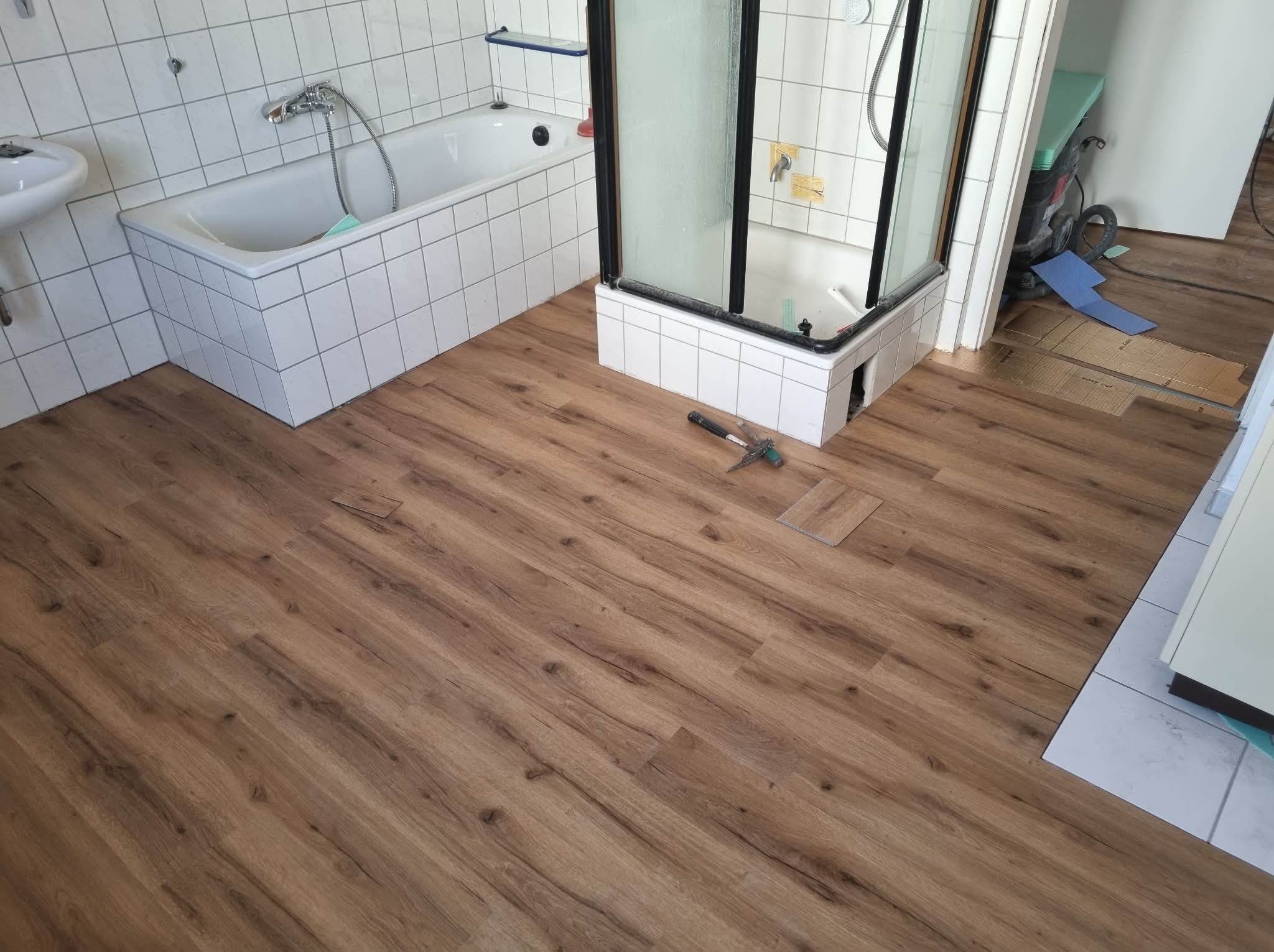 Ułożona podłoga z paneli imitujących drewno w łazience z białą wanną i kabiną prysznicową. Widoczny młotek i fragment panela. Jasne płytki na ścianach.