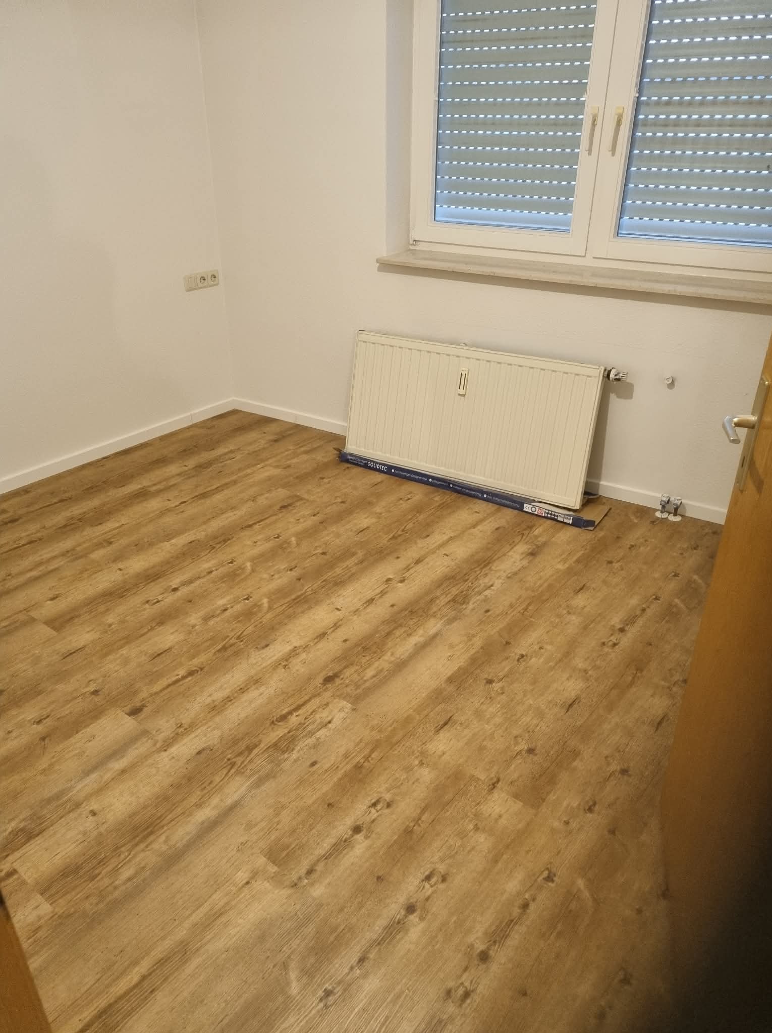 Jasne panele podłogowe w pokoju z grzejnikiem pod oknem z żaluzjami. Widoczne listwy przypodłogowe i fragment drzwi. Minimalistyczne wnętrze w neutralnej kolorystyce.