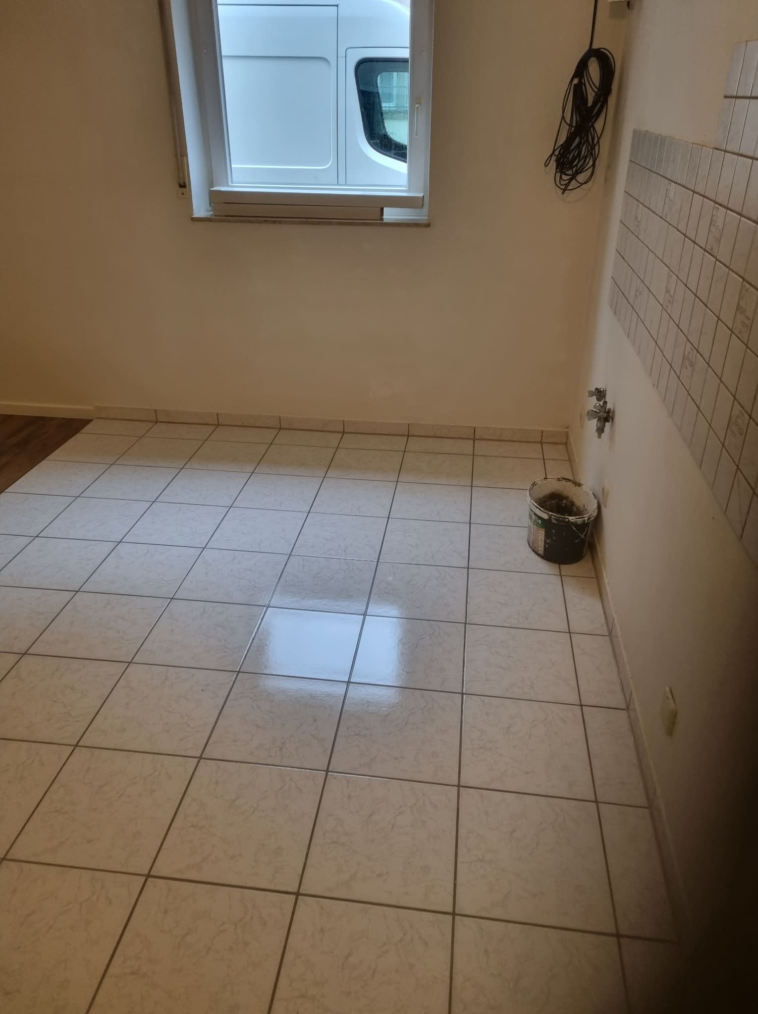 Jasne płytki podłogowe w trakcie układania, widoczny fragment ściany z częściowo położonymi płytkami, wiadro z zaprawą i okno z widokiem na samochód dostawczy.