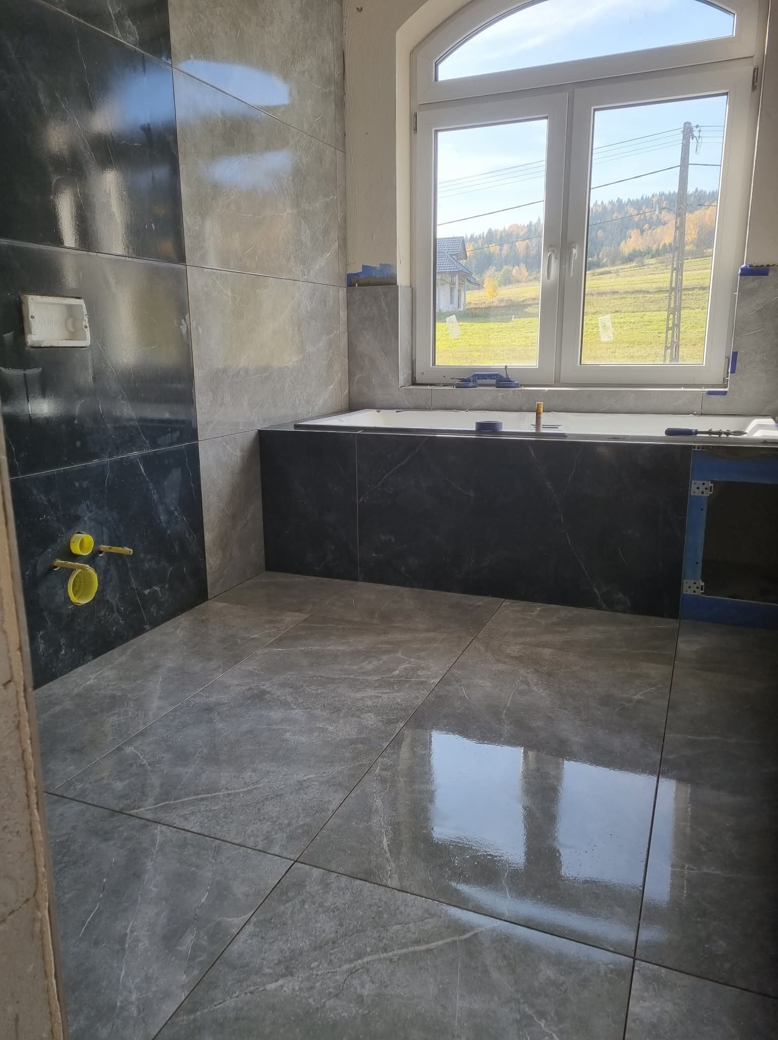 Częściowo wykończona łazienka z szarymi i czarnymi płytkami imitującymi marmur, widoczne żółte rury i okno z widokiem na krajobraz.