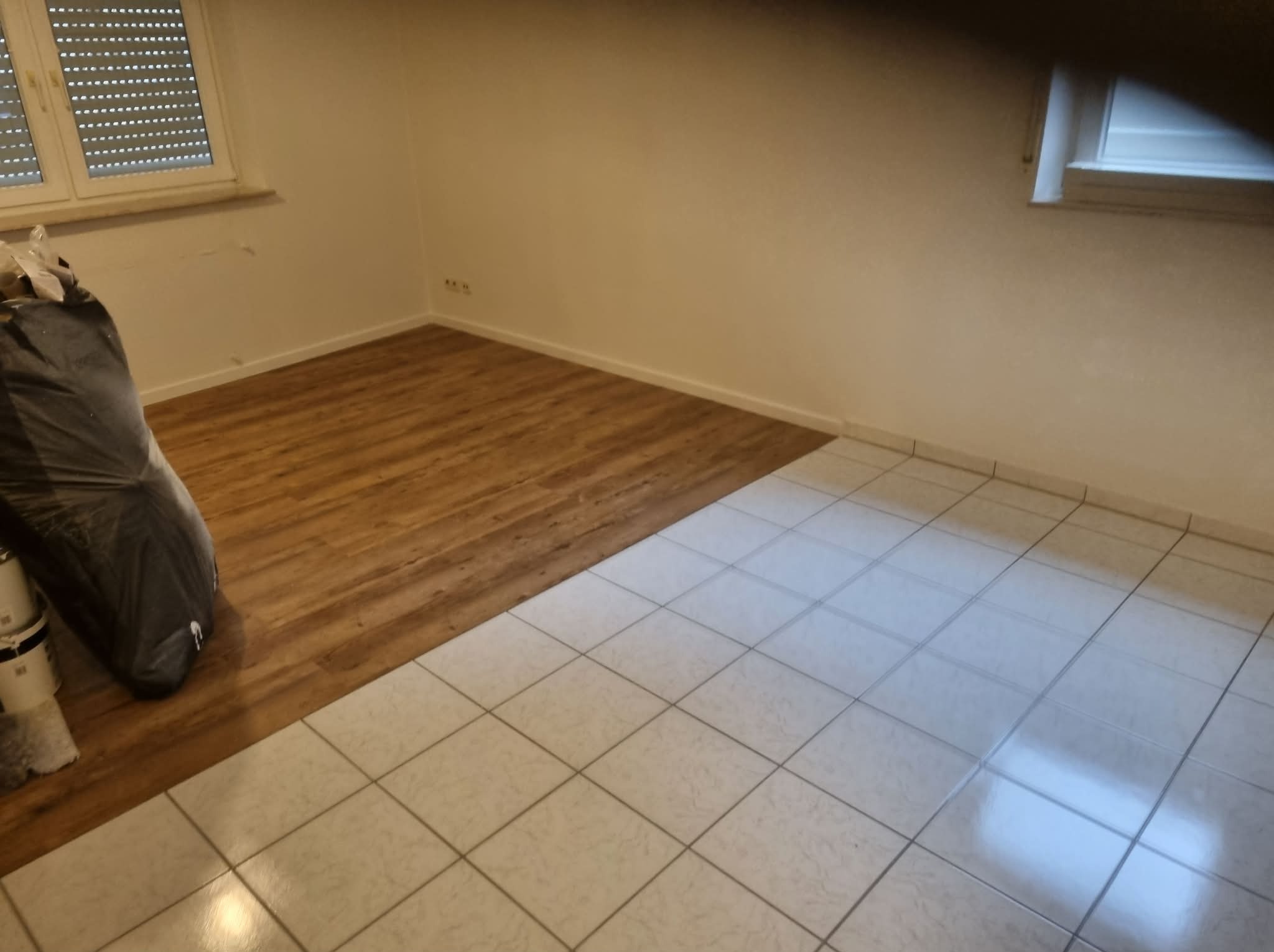 Fragment pokoju z częściowo ułożoną podłogą: panele imitujące drewno i jasne płytki. W rogu worki z materiałami budowlanymi. Widoczne okno z żaluzjami.