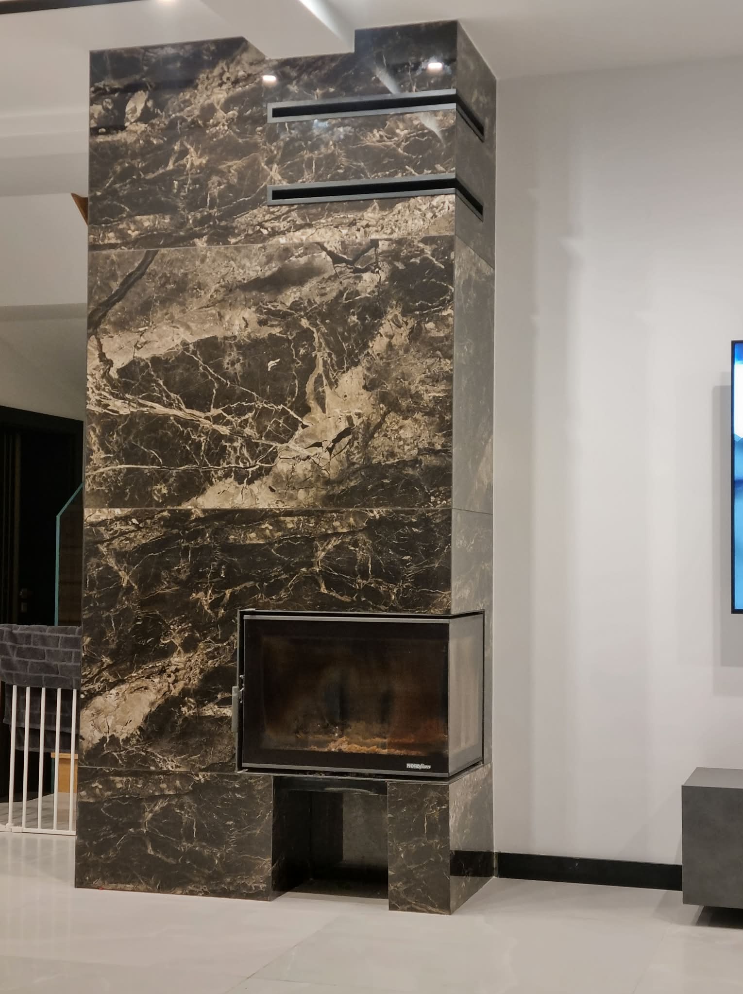 Obudowa kominka z ciemnego marmuru z wyraźnym wzorem żyłkowania, w nowoczesnym salonie z białymi ścianami i jasną podłogą. Widoczne detale wykończenia.