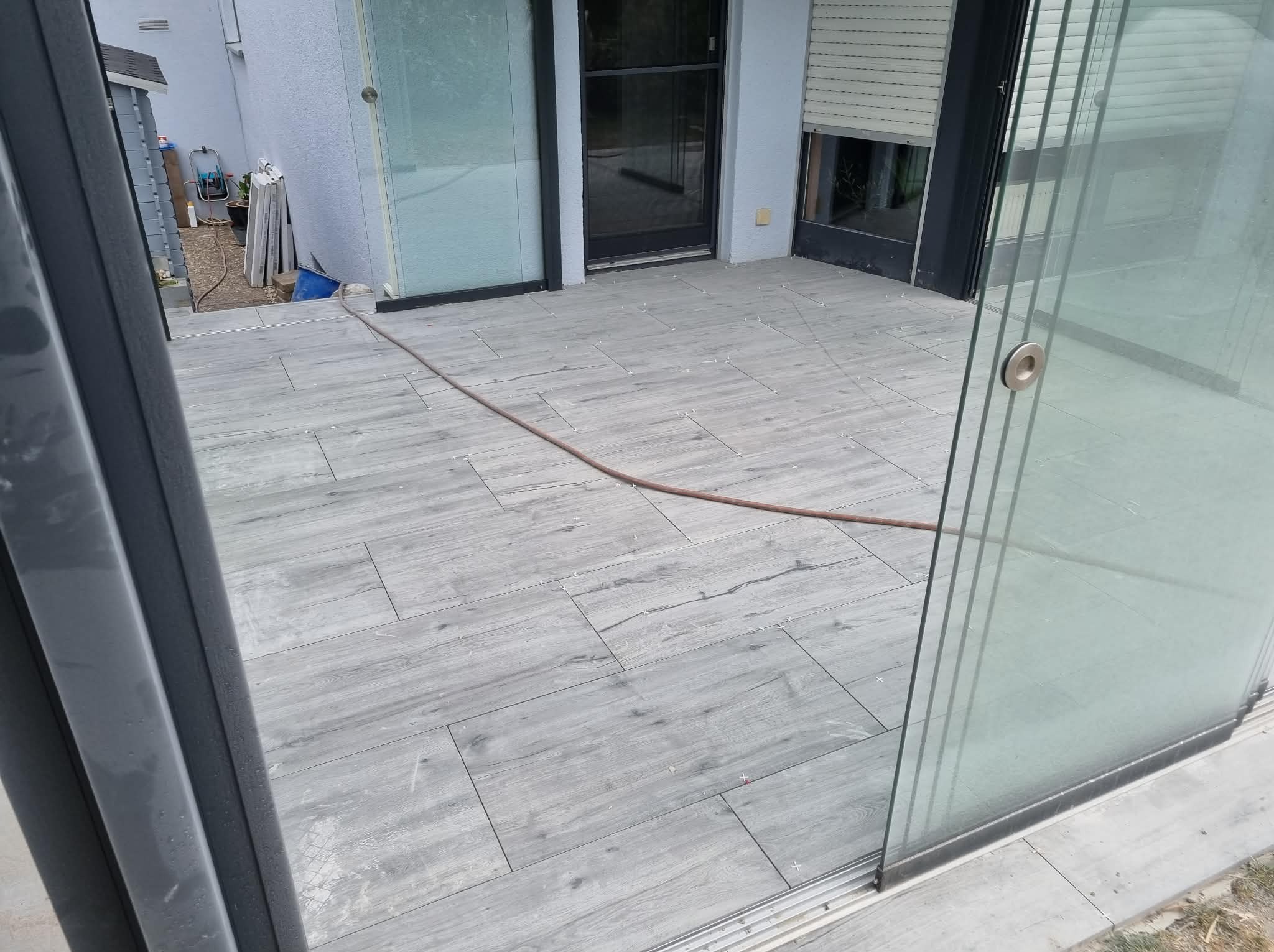 Jasnoszara, drewnopodobna terakota na balkonie z przesuwnymi drzwiami szklanymi. Widoczne krzyżyki dystansowe i wąż ogrodowy. Nowoczesne wykończenie.