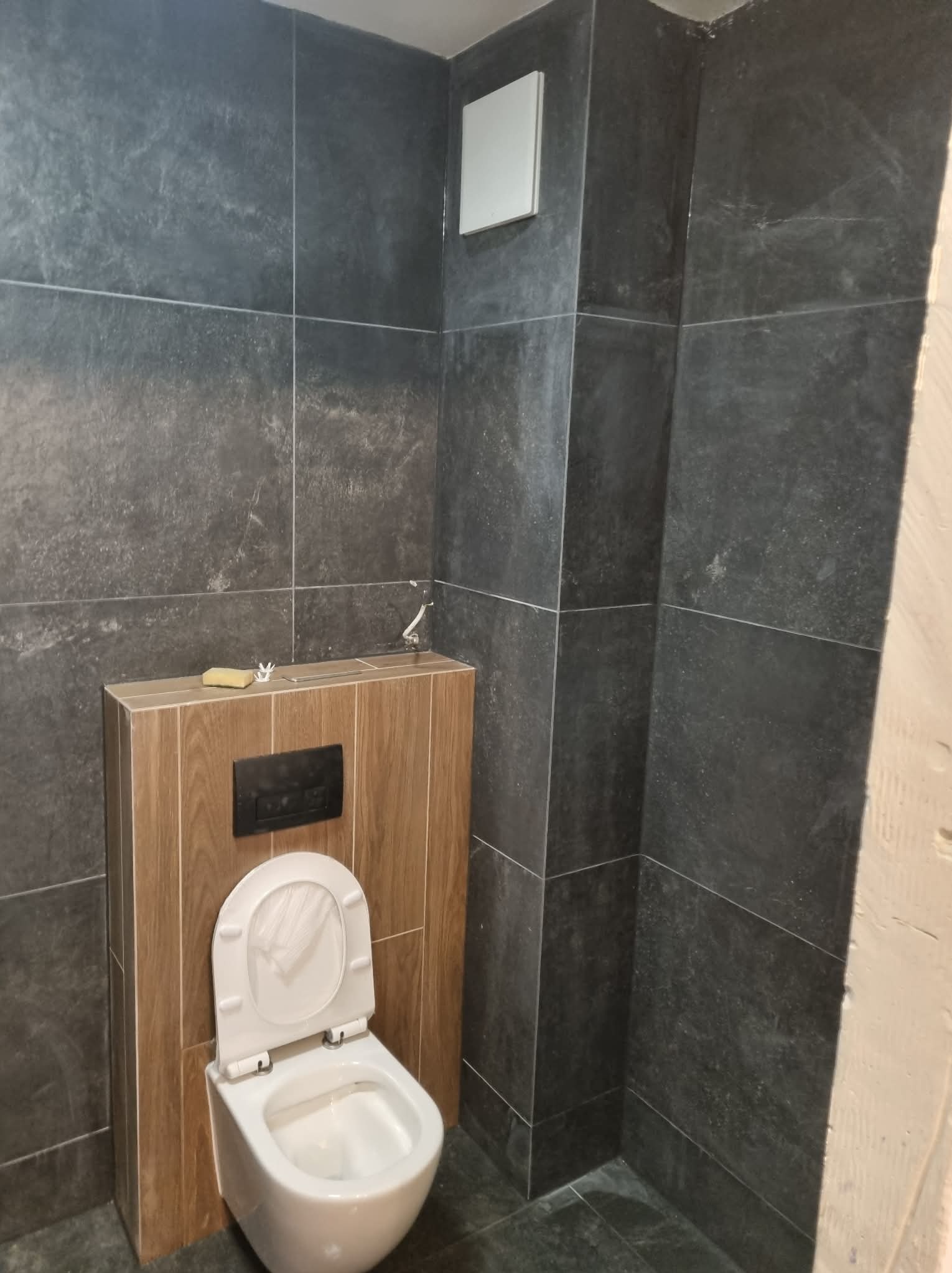 Nowoczesna toaleta podwieszana z drewnianą obudową, otwartą klapą i szarymi płytkami na ścianach. Widoczne narzędzia i gąbka na obudowie. Minimalistyczny design.