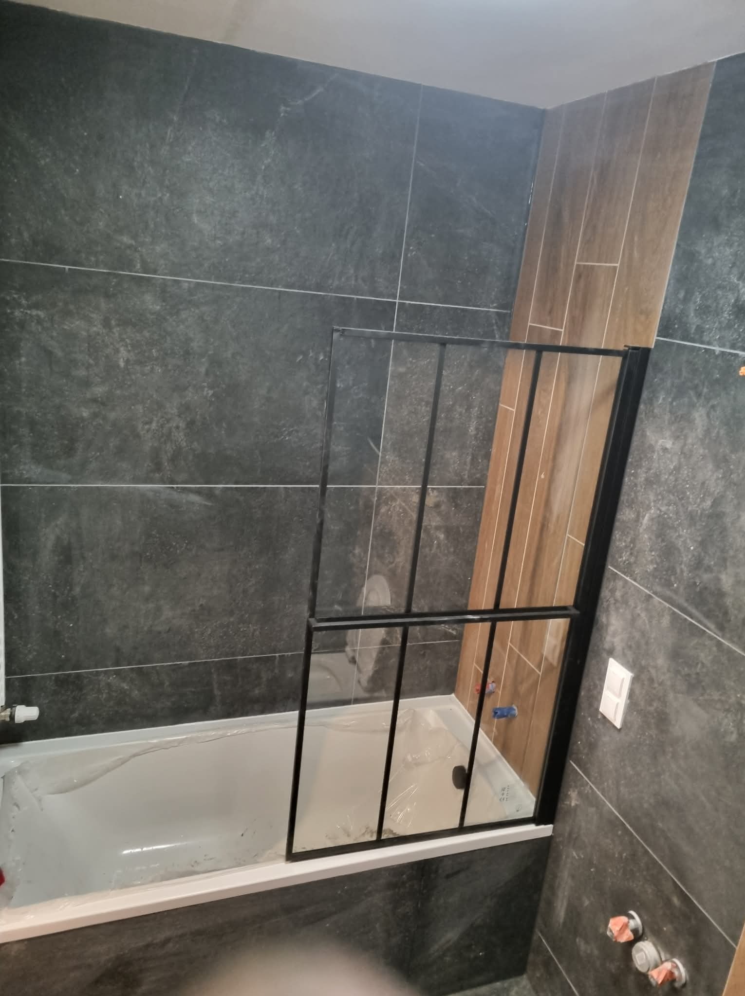 Nowoczesna łazienka w trakcie wykańczania: szara, teksturowana płytka, drewnopodobna okładzina, biała wanna z czarną, minimalistyczną kabiną prysznicową w folii ochronnej.