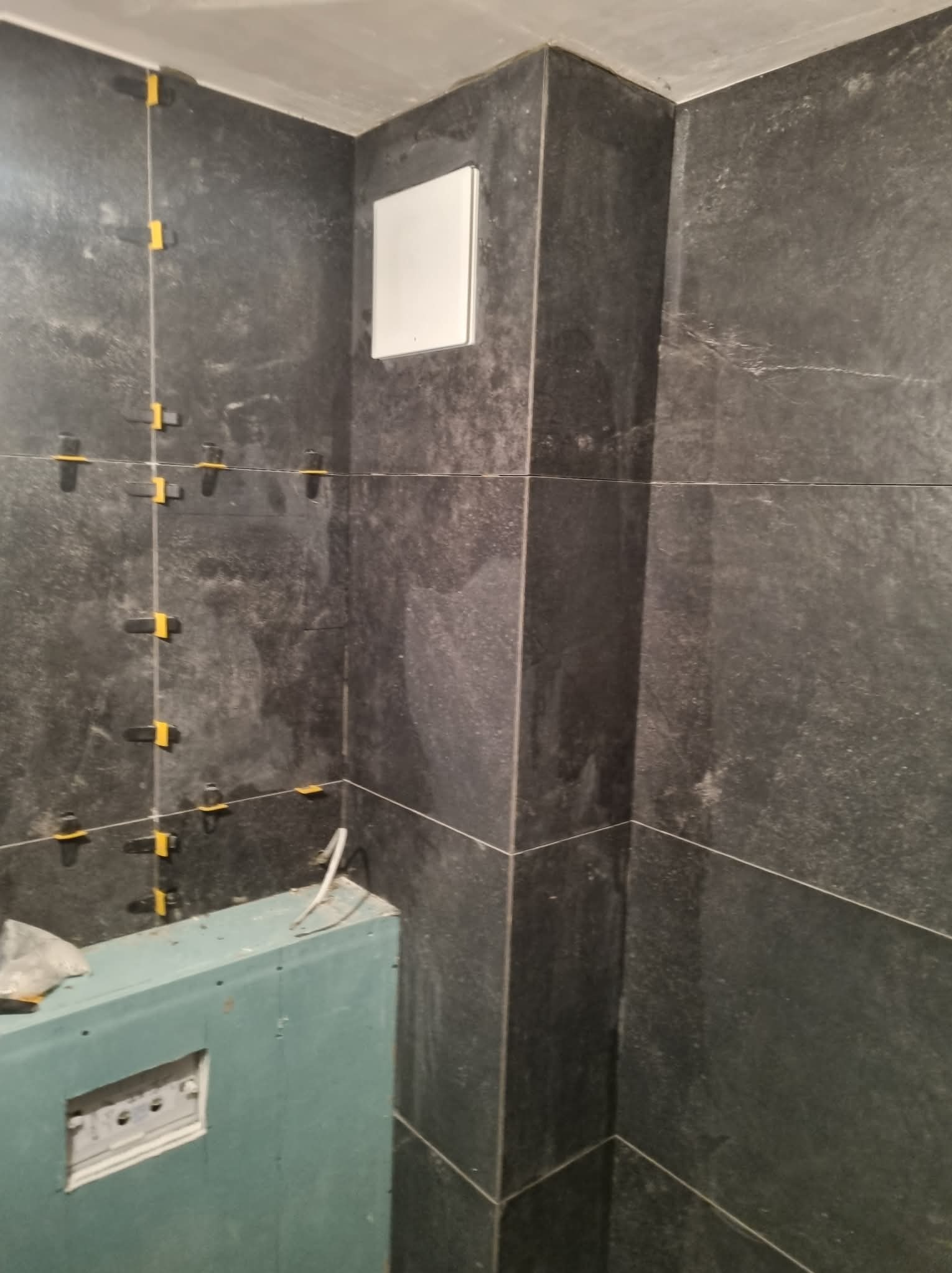 Ciemne, matowe płytki na ścianie w trakcie montażu, z widocznymi krzyżykami dystansowymi. Fragment zabudowy z płyty gipsowej w dolnej części kadru.