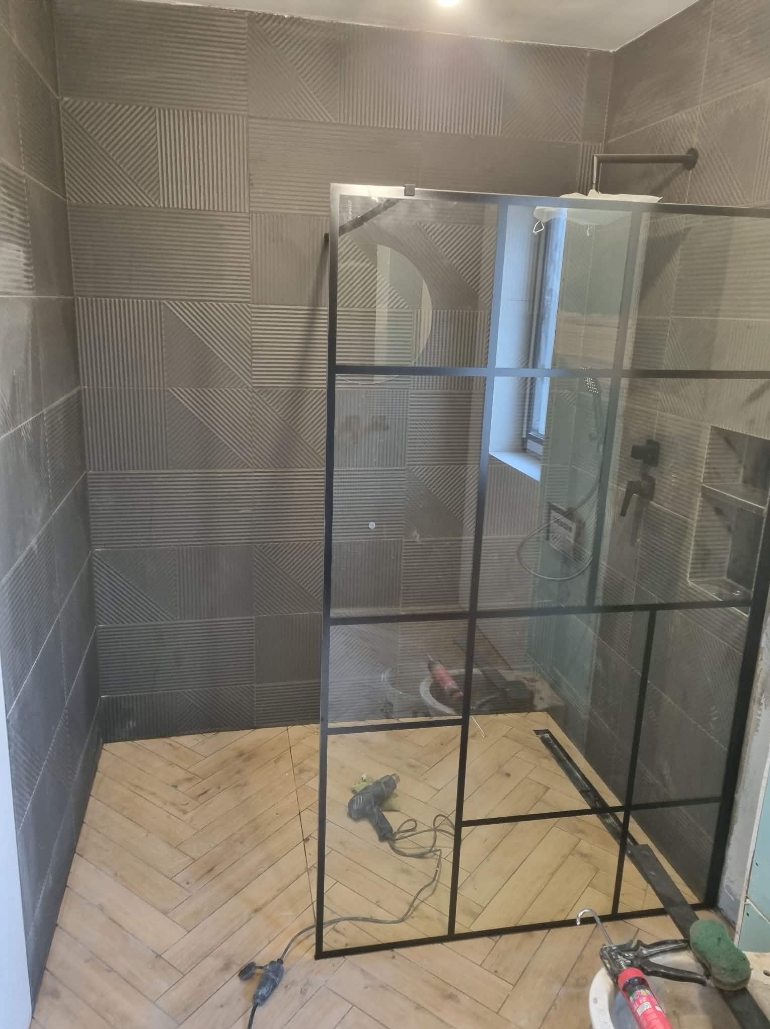 Nowoczesna łazienka w trakcie wykańczania: szare płytki z wzorem, drewnopodobna podłoga w jodełkę, czarna kabina prysznicowa i narzędzia glazurnicze.