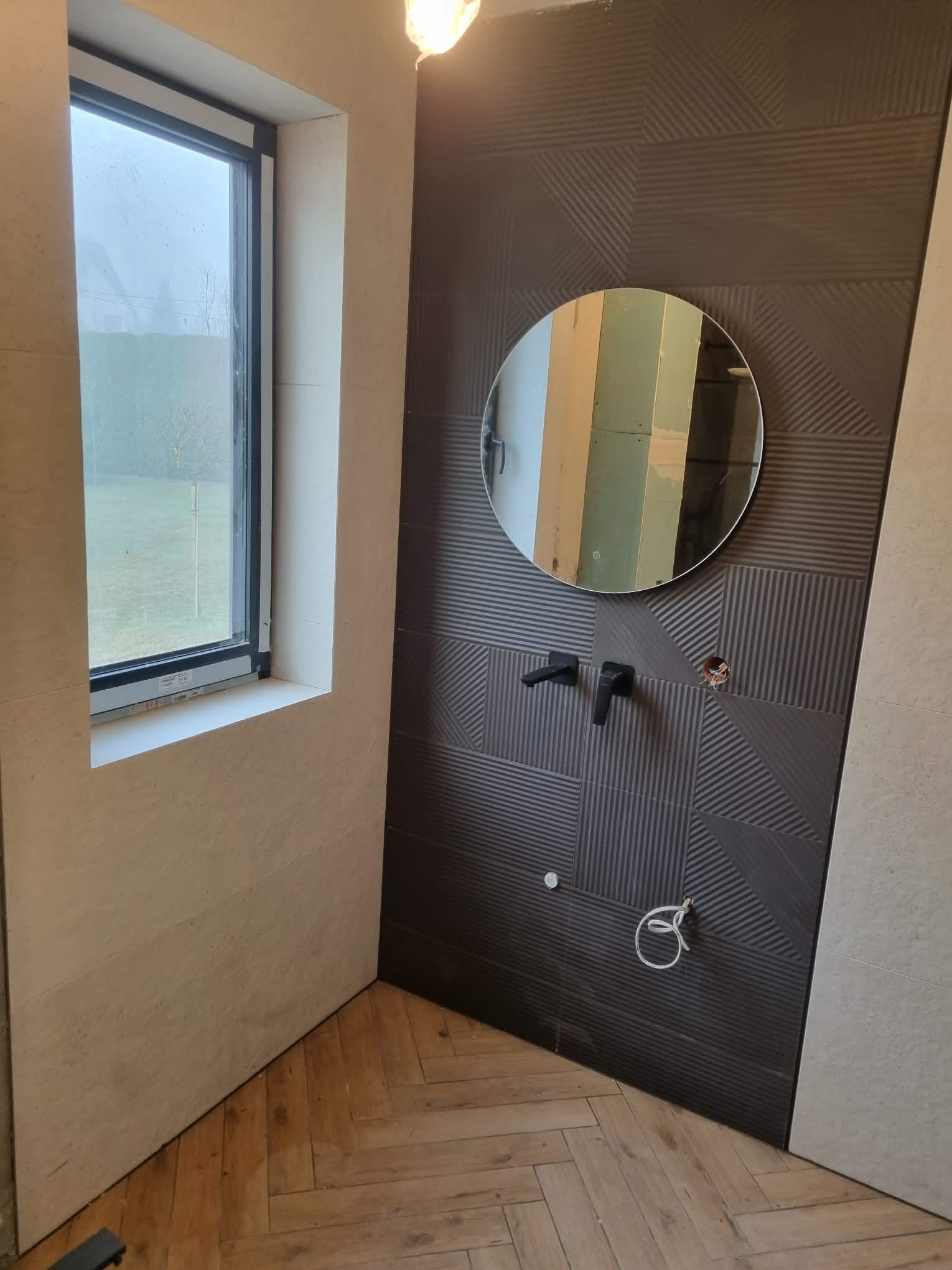 Nowoczesna łazienka w trakcie wykańczania: ciemne, wzorzyste płytki, okrągłe lustro, drewniana podłoga w jodełkę, widoczne elementy instalacji. Minimalistyczny design.