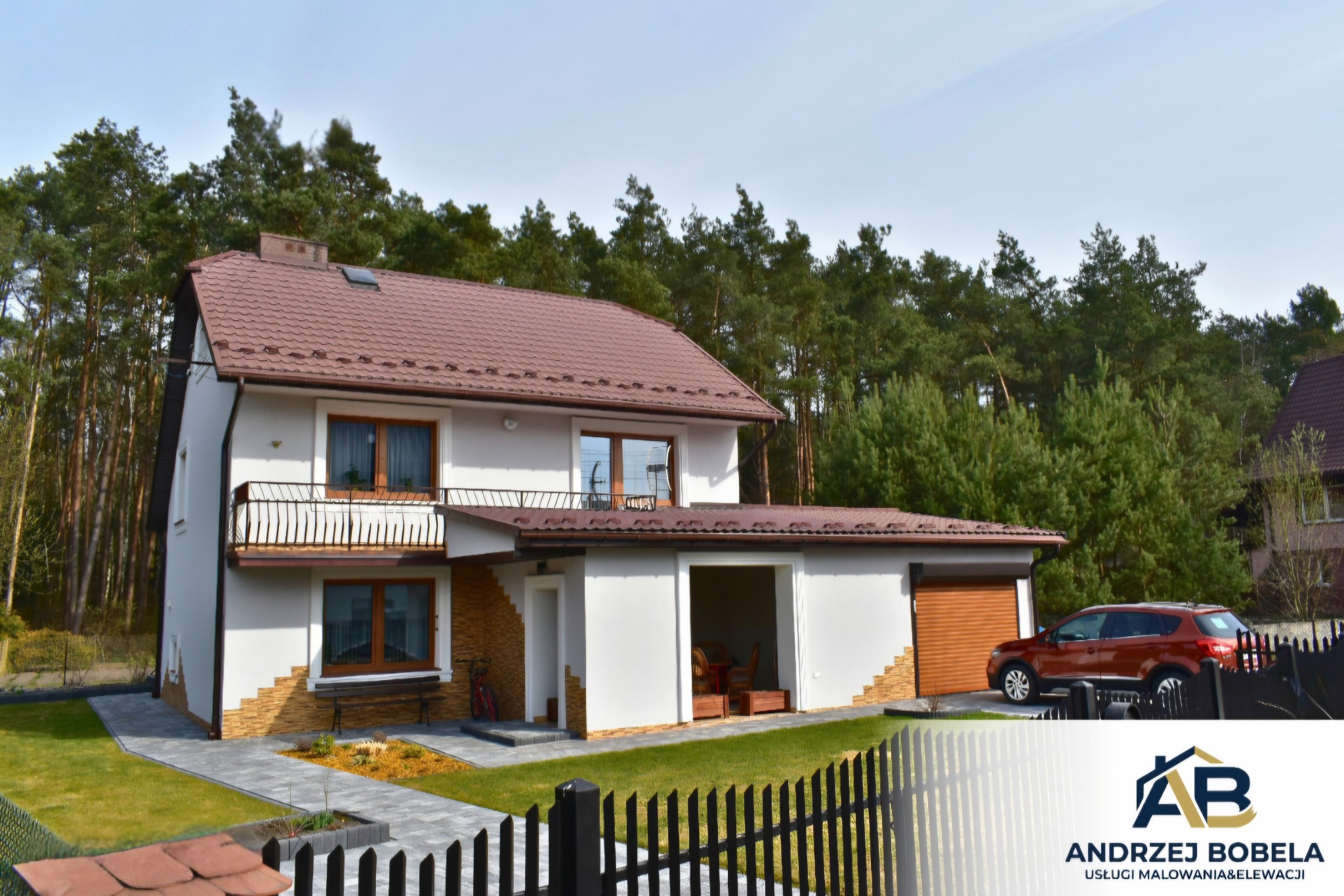 Dwukondygnacyjny dom z białymi ścianami i brązowym dachem, otoczony zielenią, z widocznym fragmentem ogrodzenia i samochodem. Ukończona realizacja.