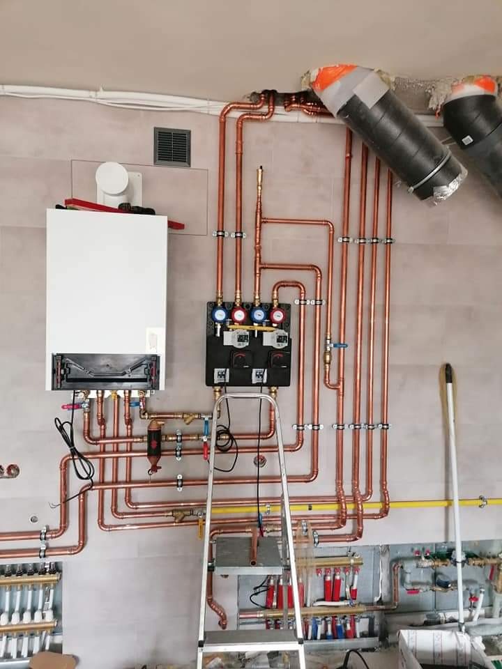 Biały piec gazowy z miedzianą instalacją, czarna skrzynka sterująca, drabina. Widoczne rury i zawory. Instalacja grzewcza w trakcie montażu, surowy stan ścian.